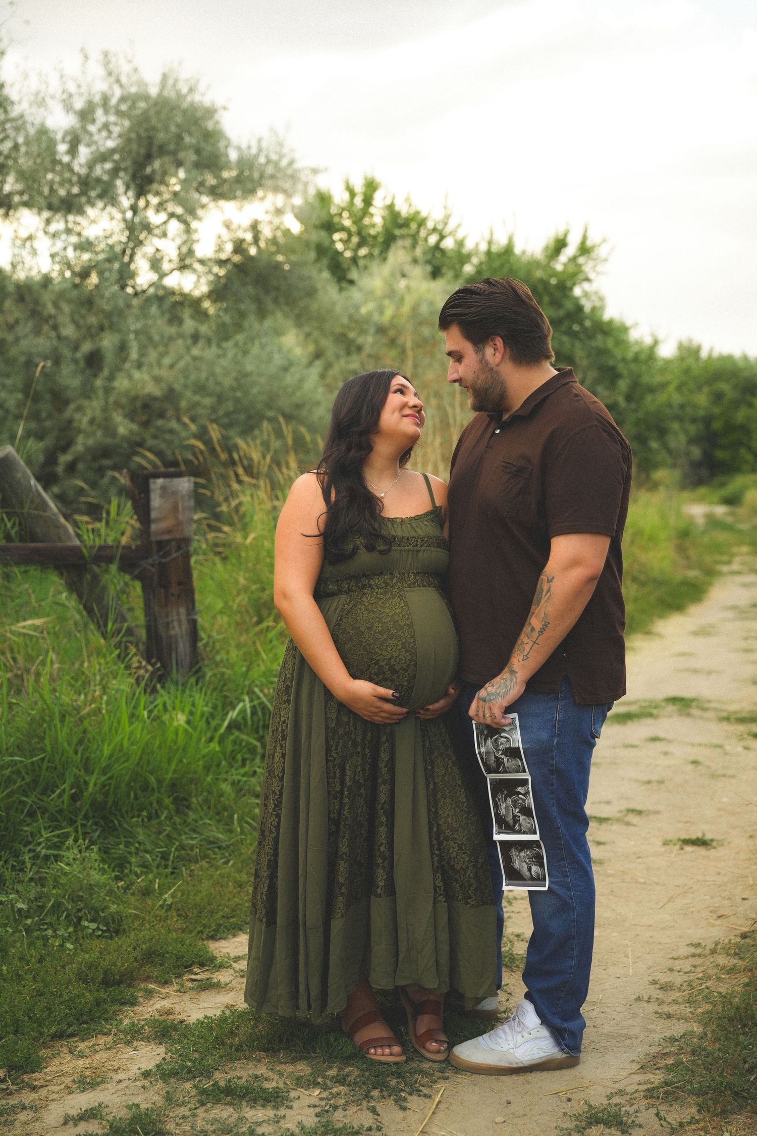 Pregnant couple holding ultrasound photos on a garden path, woman in green maxi dress, man in brown shirt and jeans.