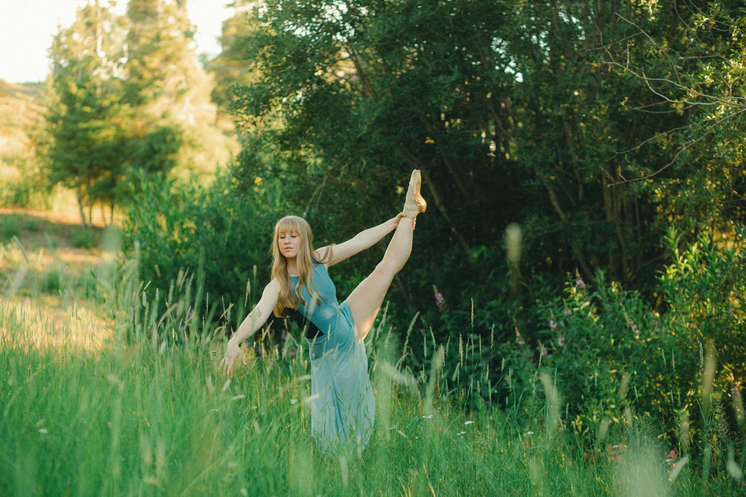 A flexible woman in a teal dress performs a standing split in a lush green meadow surrounded by trees.