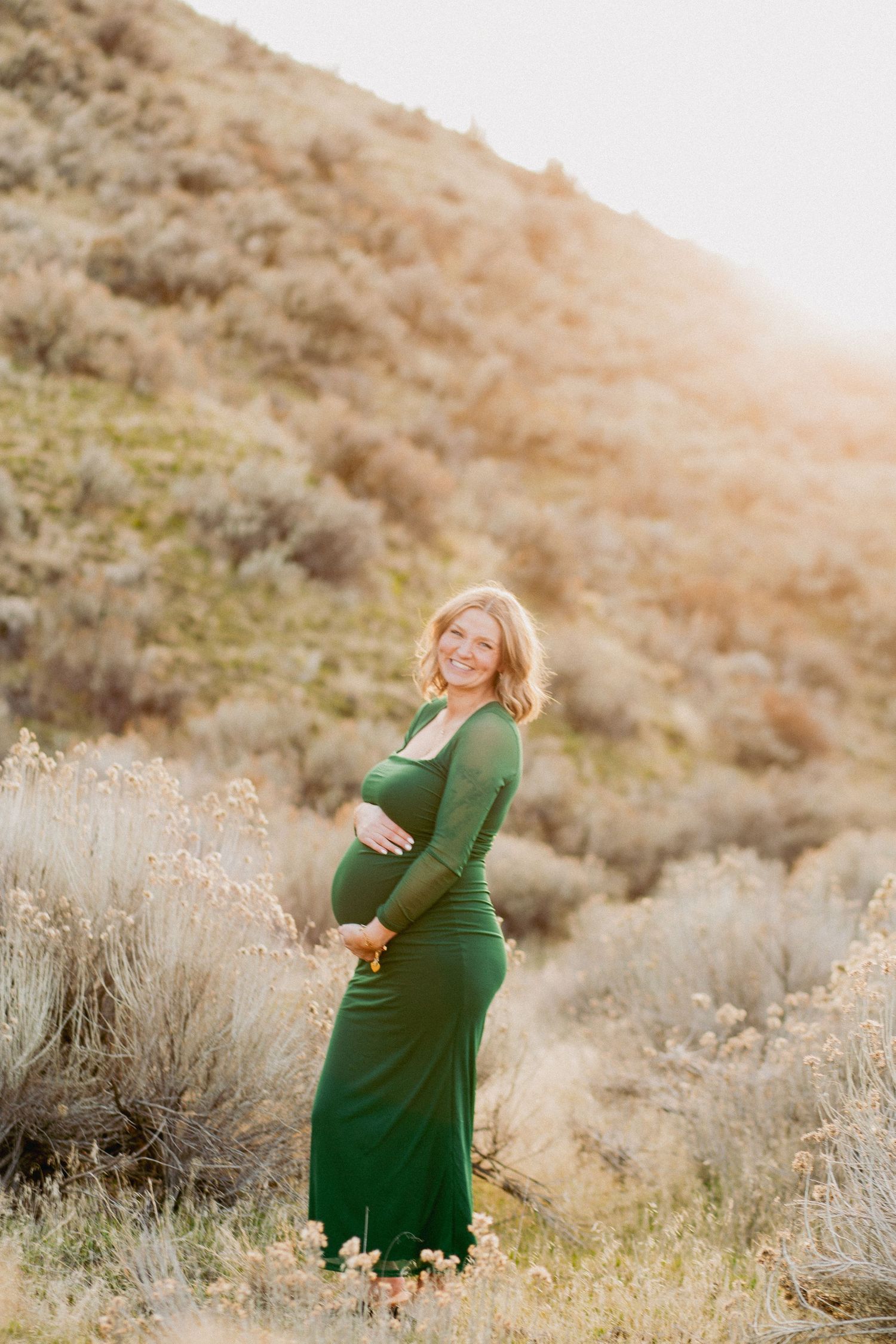 Smiling pregnant woman in green maxi dress poses outdoors in sunlit hillside field with sagebrush.