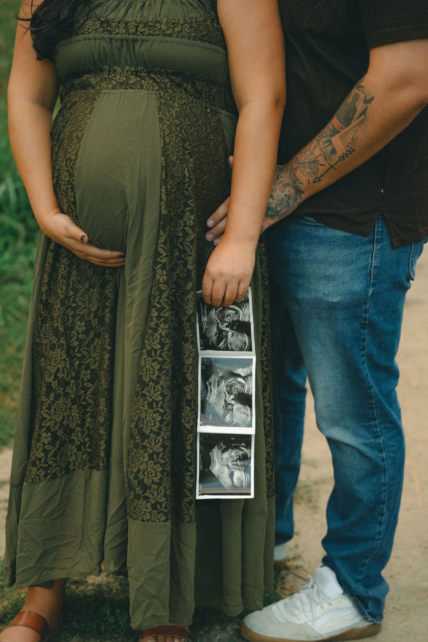 Pregnant woman in olive dress holding ultrasound photos beside tattooed partner outdoors.