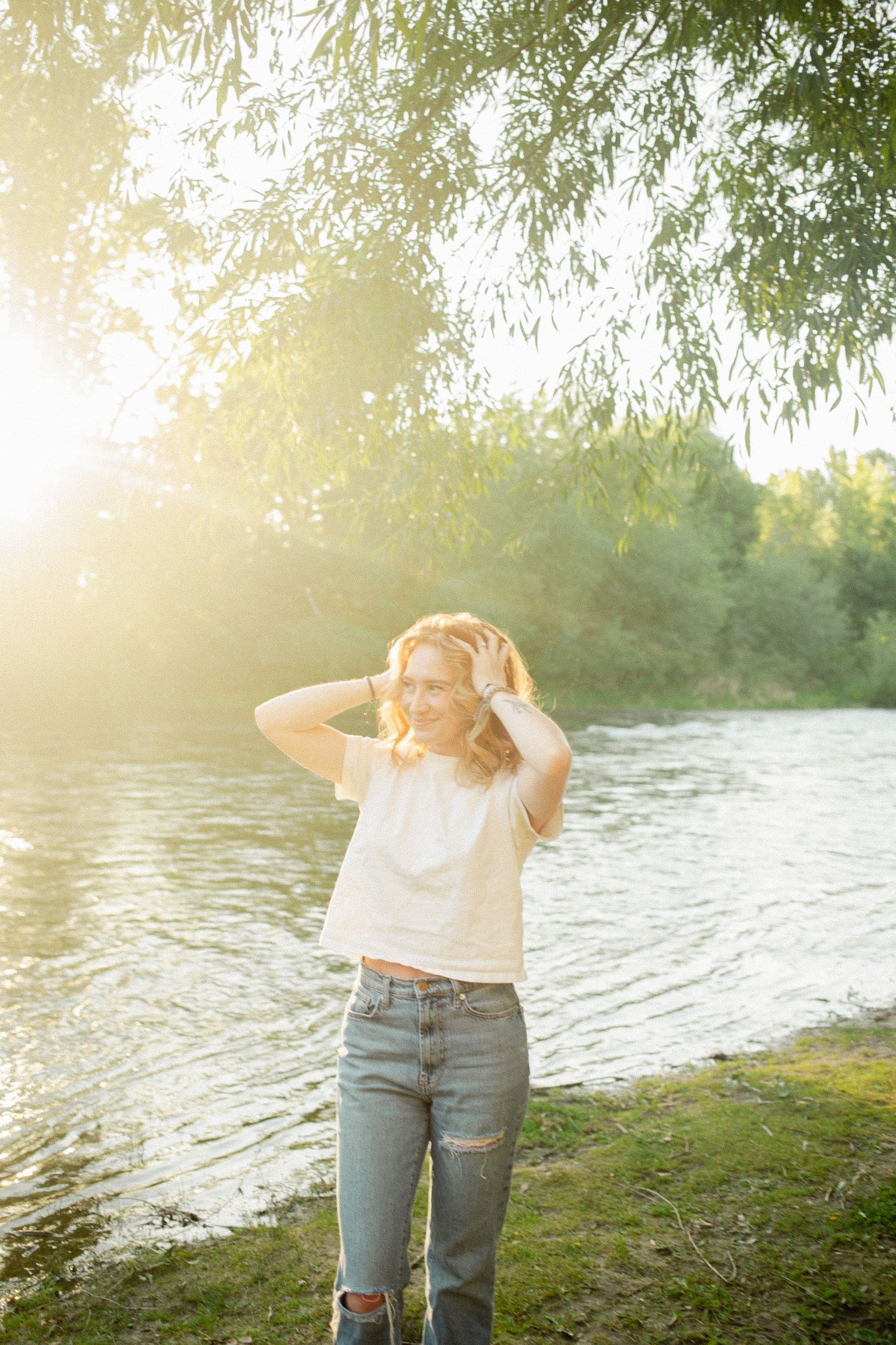Smiling woman in white crop top and jeans poses by a glowing sunlit riverside with green trees.