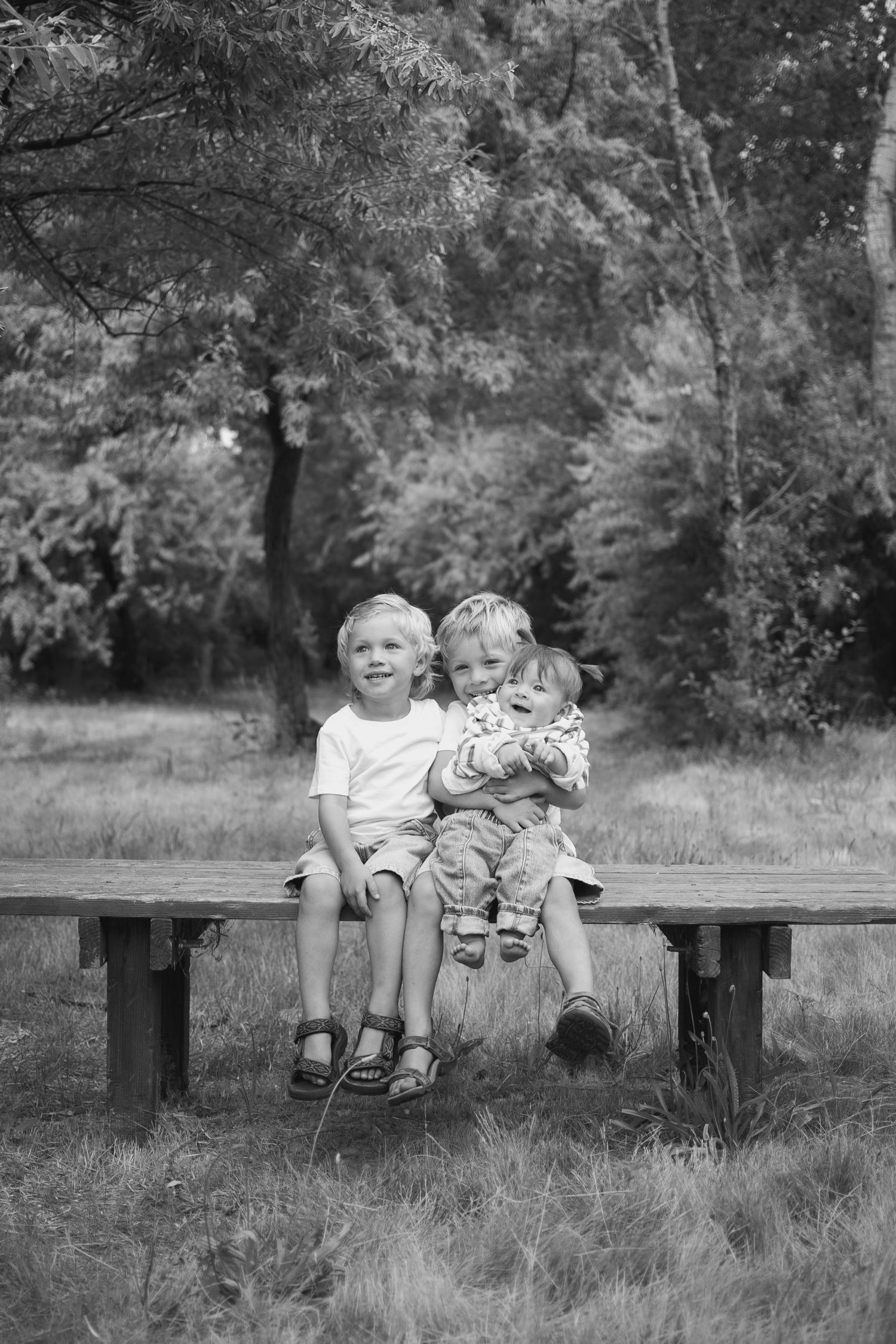 Three young siblings sit closely together on a wooden bench in a black and white photo.