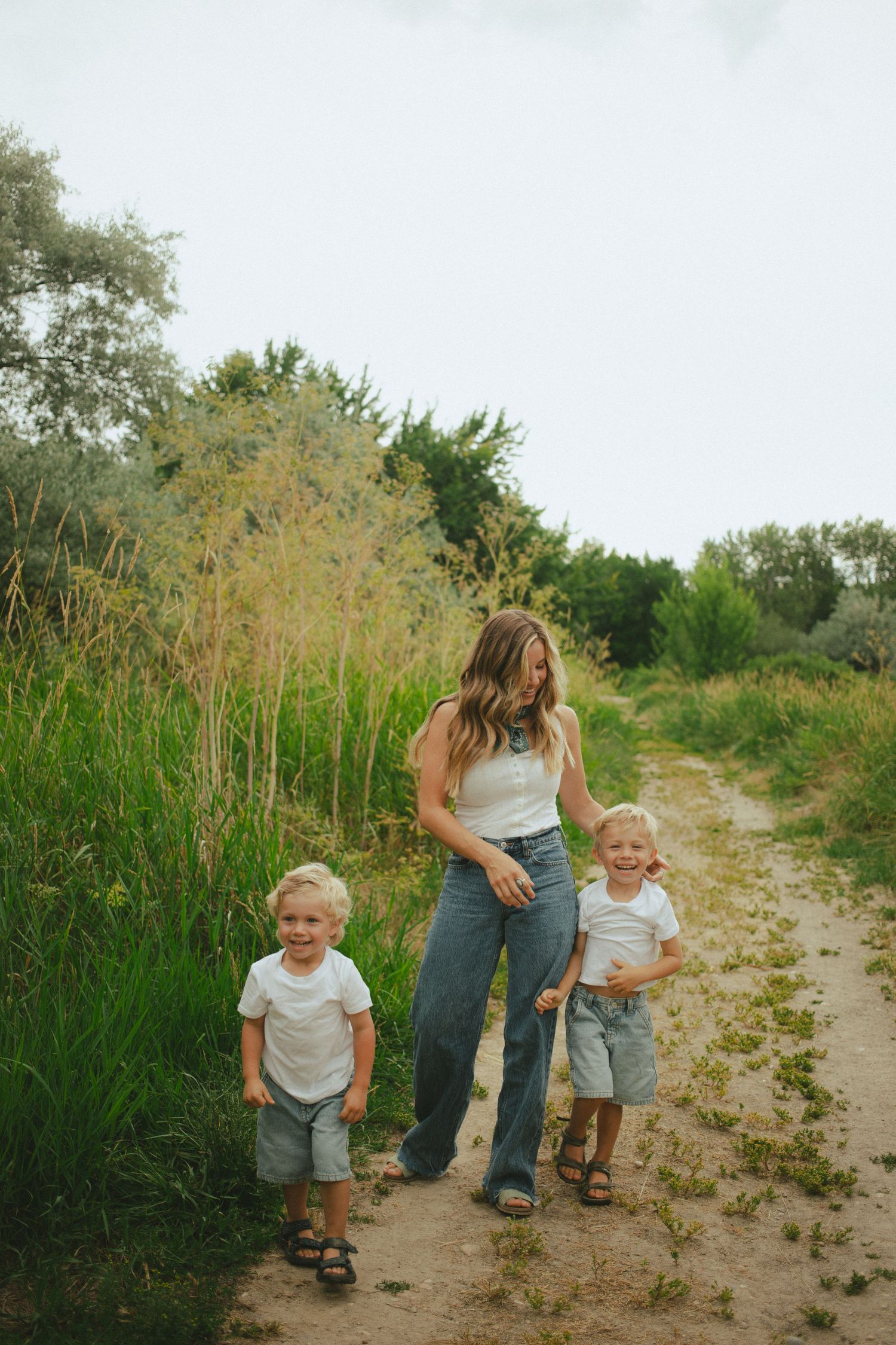 Mother in white tank and jeans walks outdoor dirt path with two young blonde toddler boys in matching outfits.