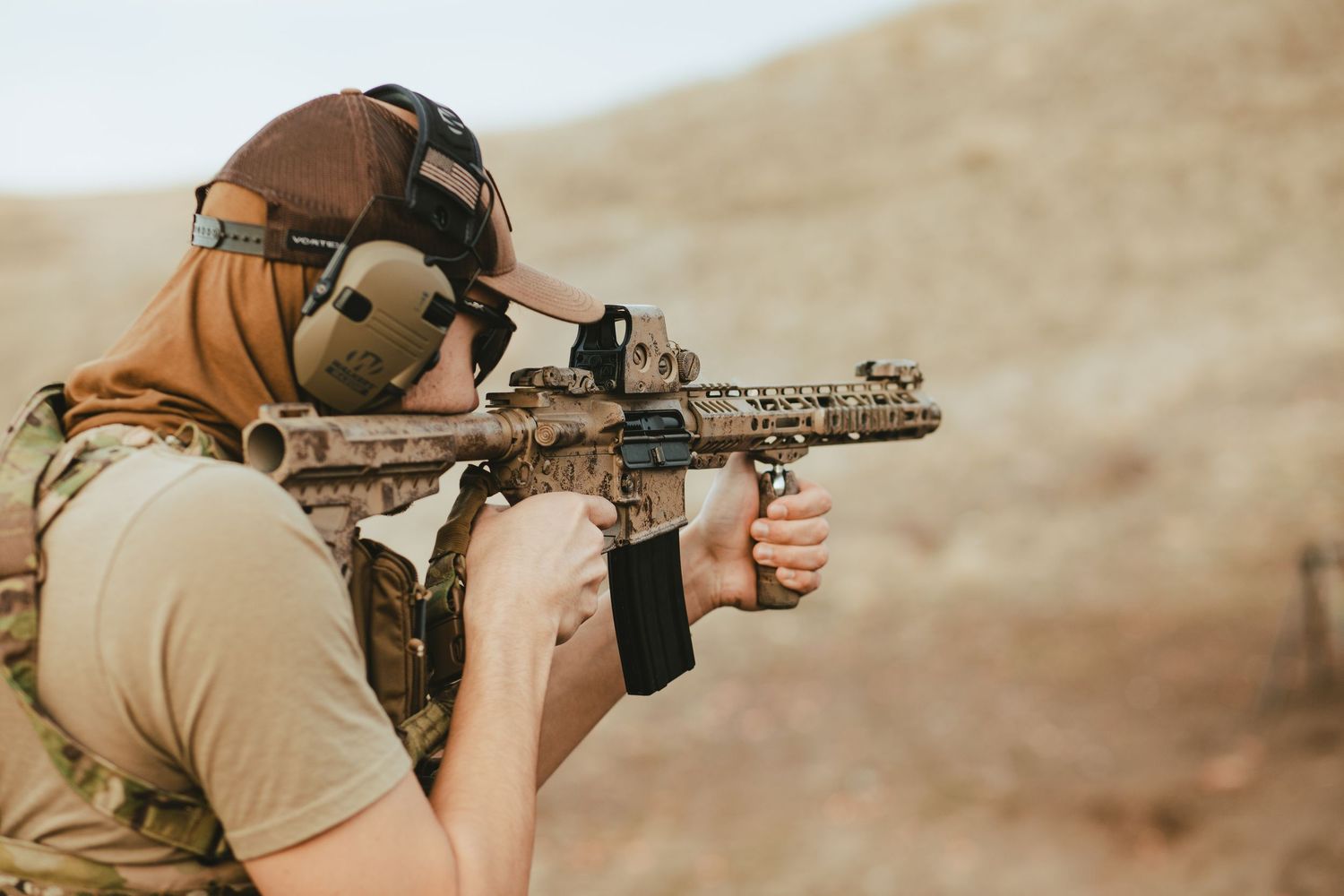 A person in tactical gear aims a rifle in a desert environment during weapons training.