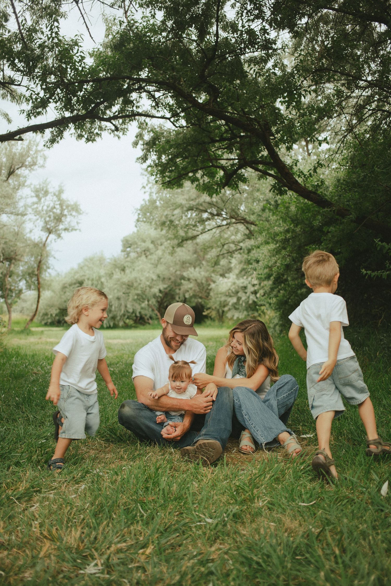 Family of five sitting on grass under trees, parents with newborn baby while two young boys play around them.