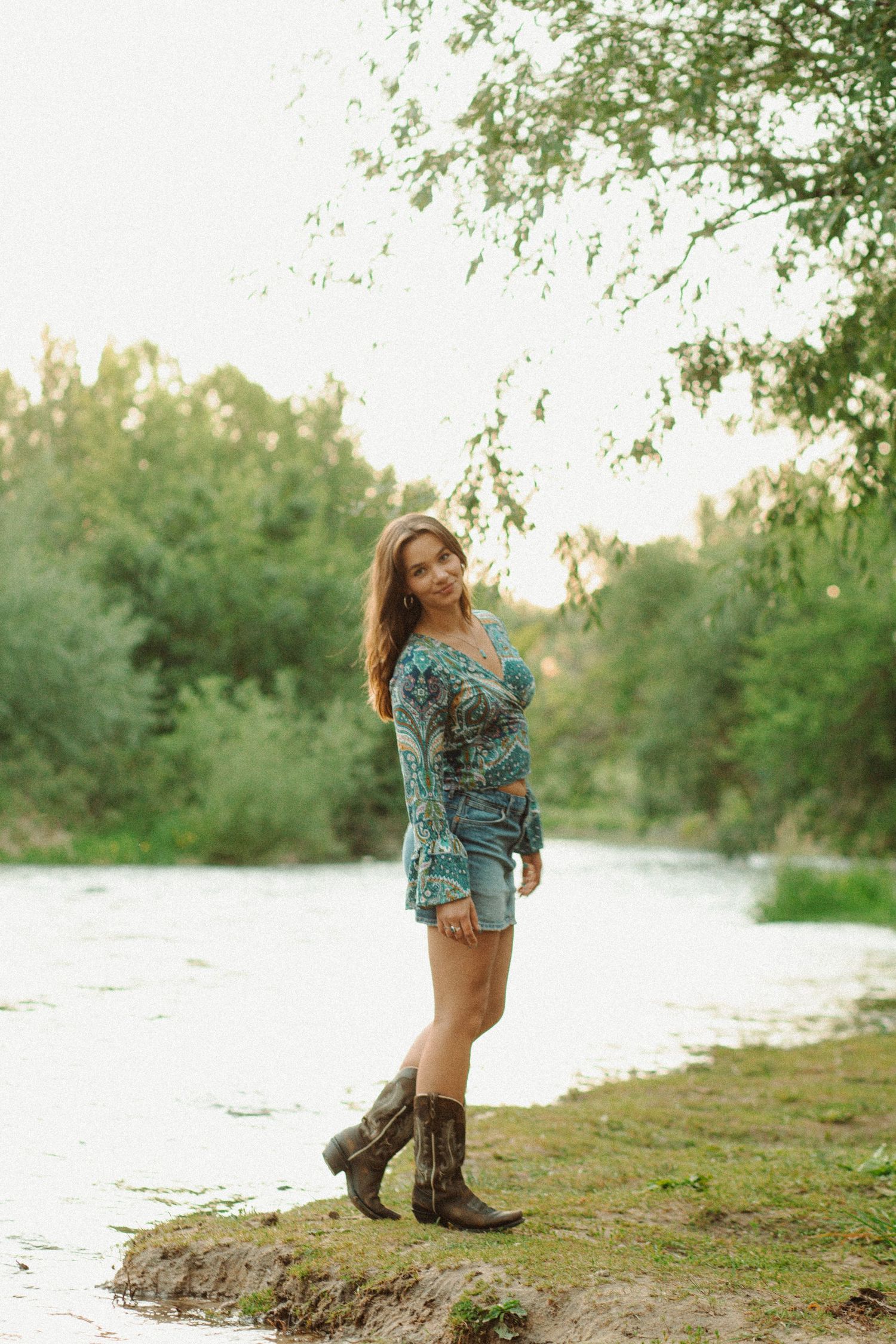 Woman in teal boho blouse, denim mini skirt, and cowboy boots smiles by a lush green riverside.