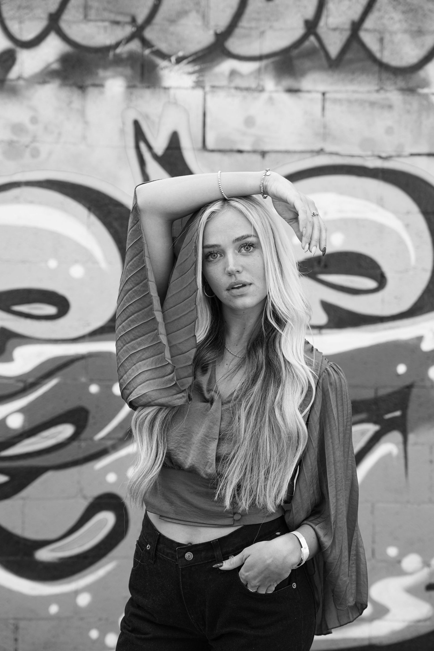 Black and white portrait of a young woman with long blonde hair posing against a graffiti wall in urban setting.