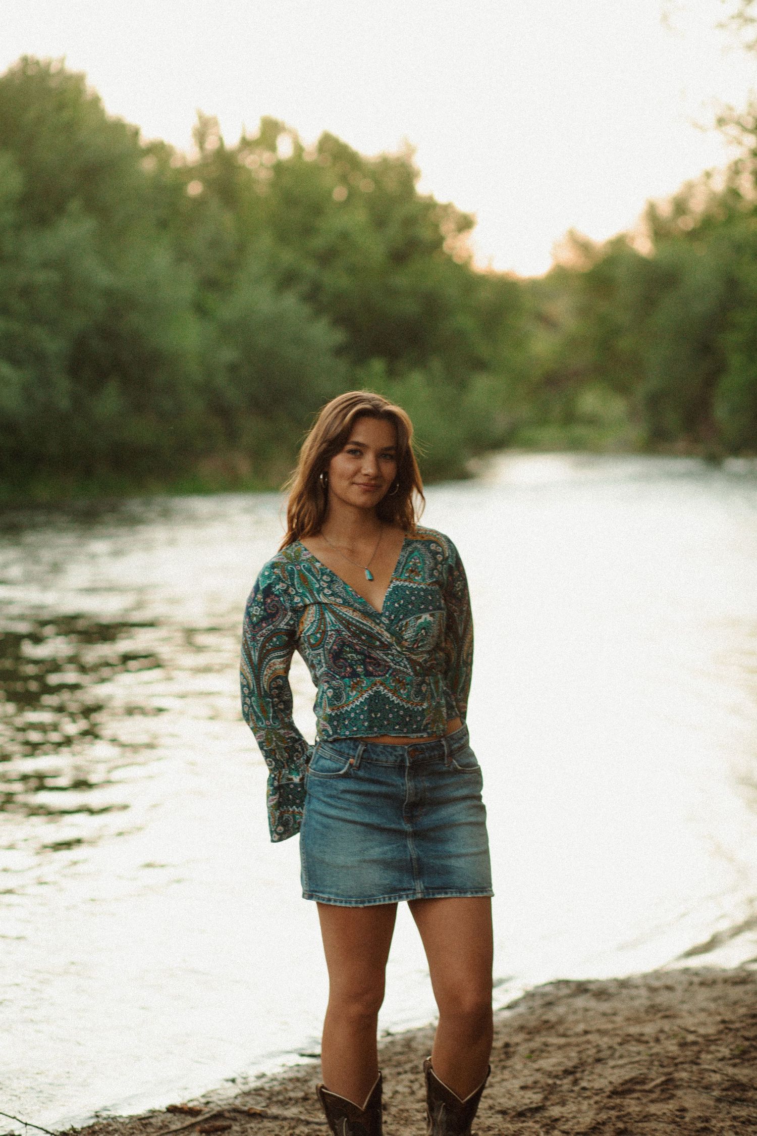 Smiling woman in boho paisley blouse and denim skirt with cowboy boots poses by a green riverbank.