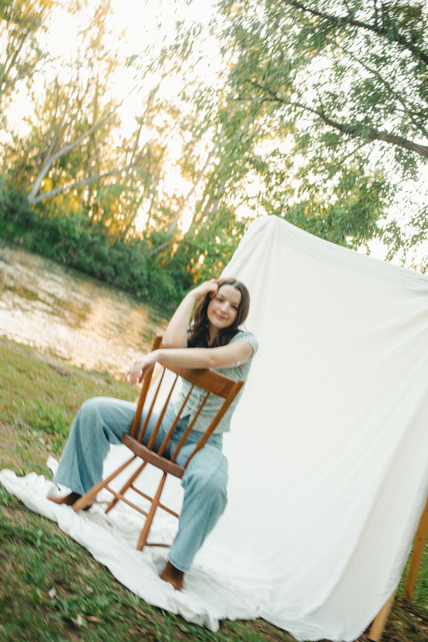 Smiling woman in blue jeans sitting on a wooden chair outdoors near a white backdrop by a sunlit river.
