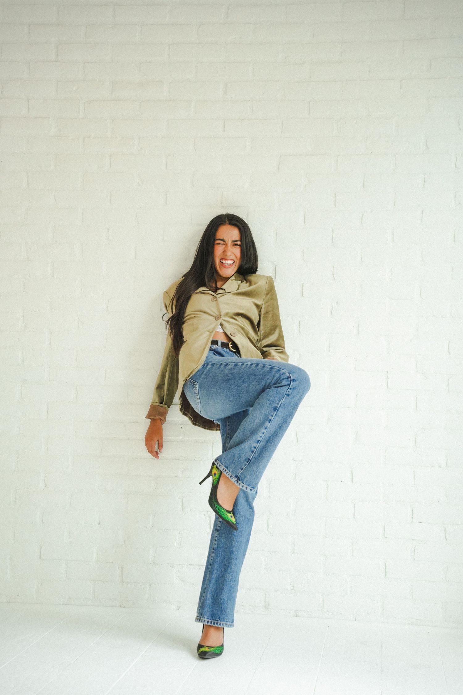 Woman in olive shirt, blue jeans, and green heels posing playfully against a white brick wall.