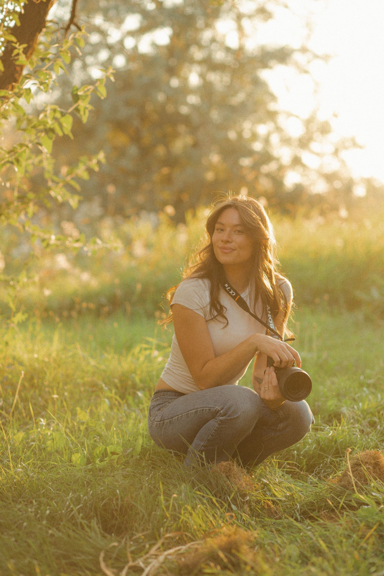 Young woman photographer crouching in sunlit meadow holding camera, wearing white tee and jeans, golden hour light filtering through trees.