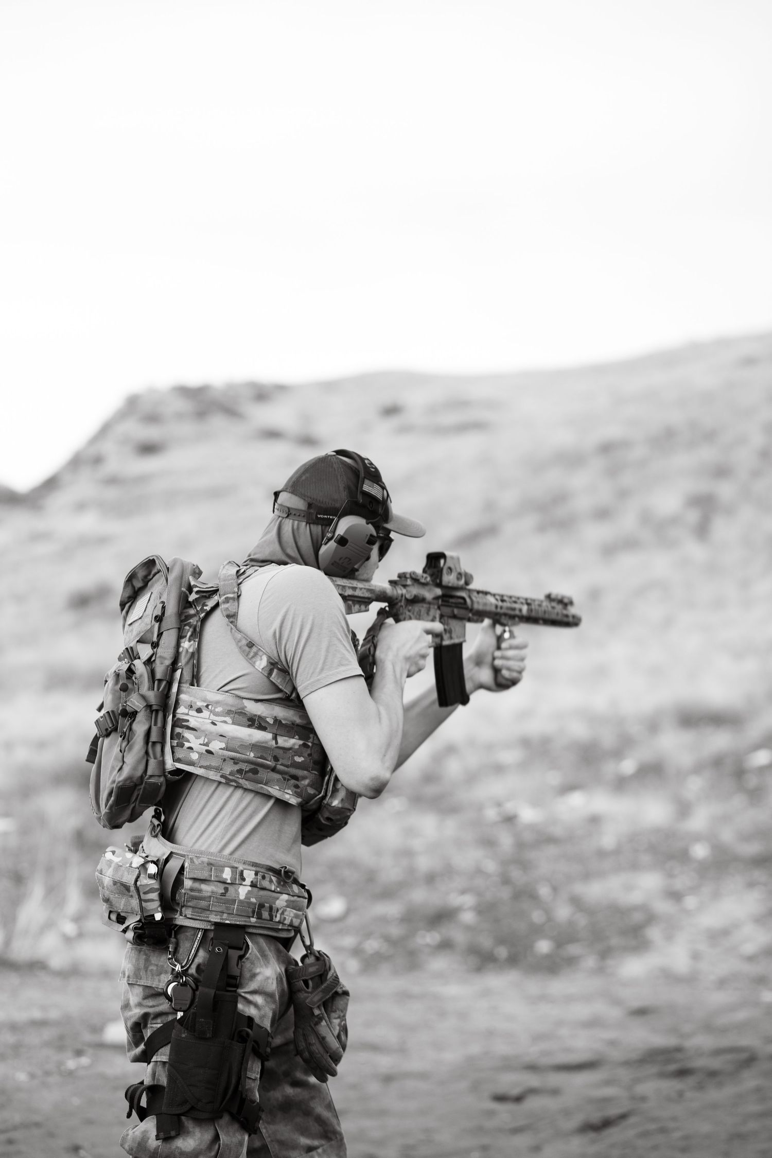 Military operator in tactical gear aims rifle while training in desert terrain, black and white photograph.