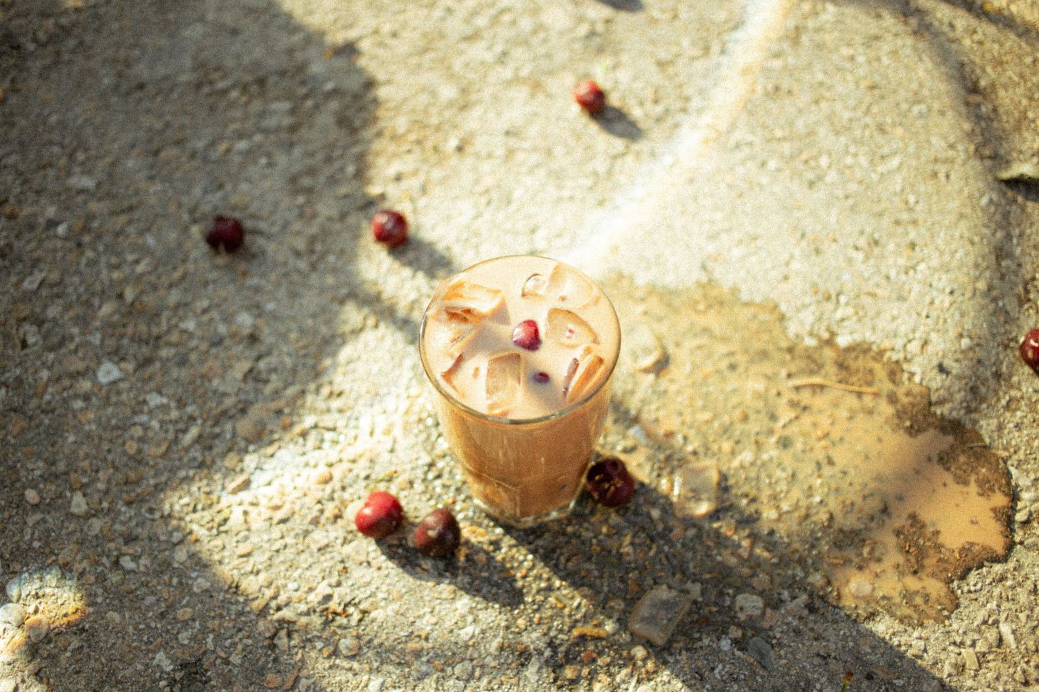 Iced coffee drink in a glass on a sunlit concrete surface surrounded by scattered red cherries.