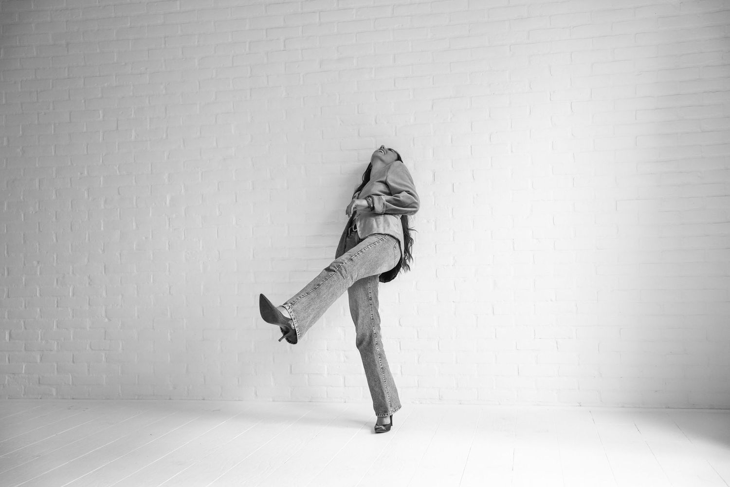 Black and white photo of a person leaning against a white brick wall with one leg raised, wearing jeans and a jacket.