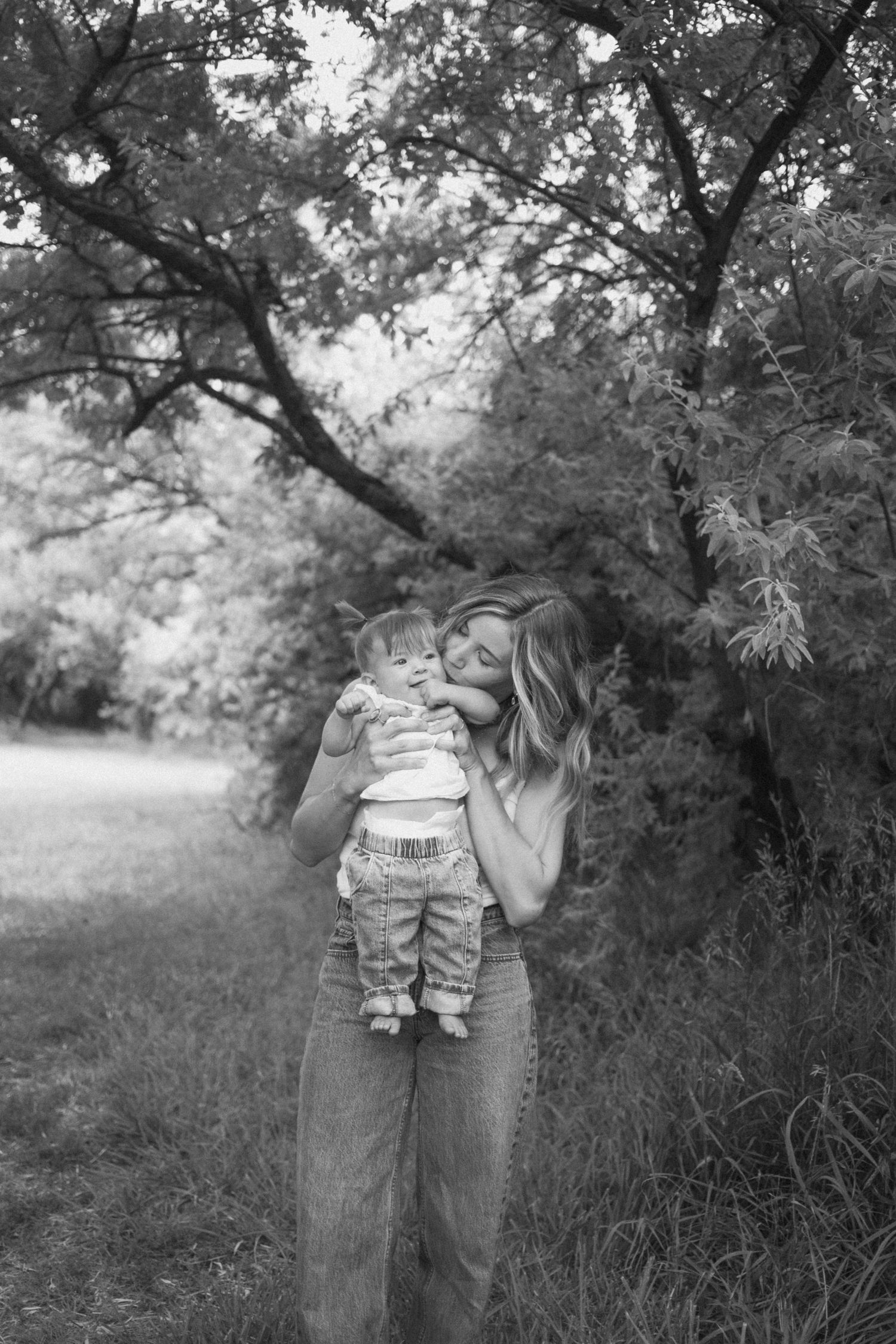 Black and white portrait of a mother tenderly kissing her baby outdoors near trees.