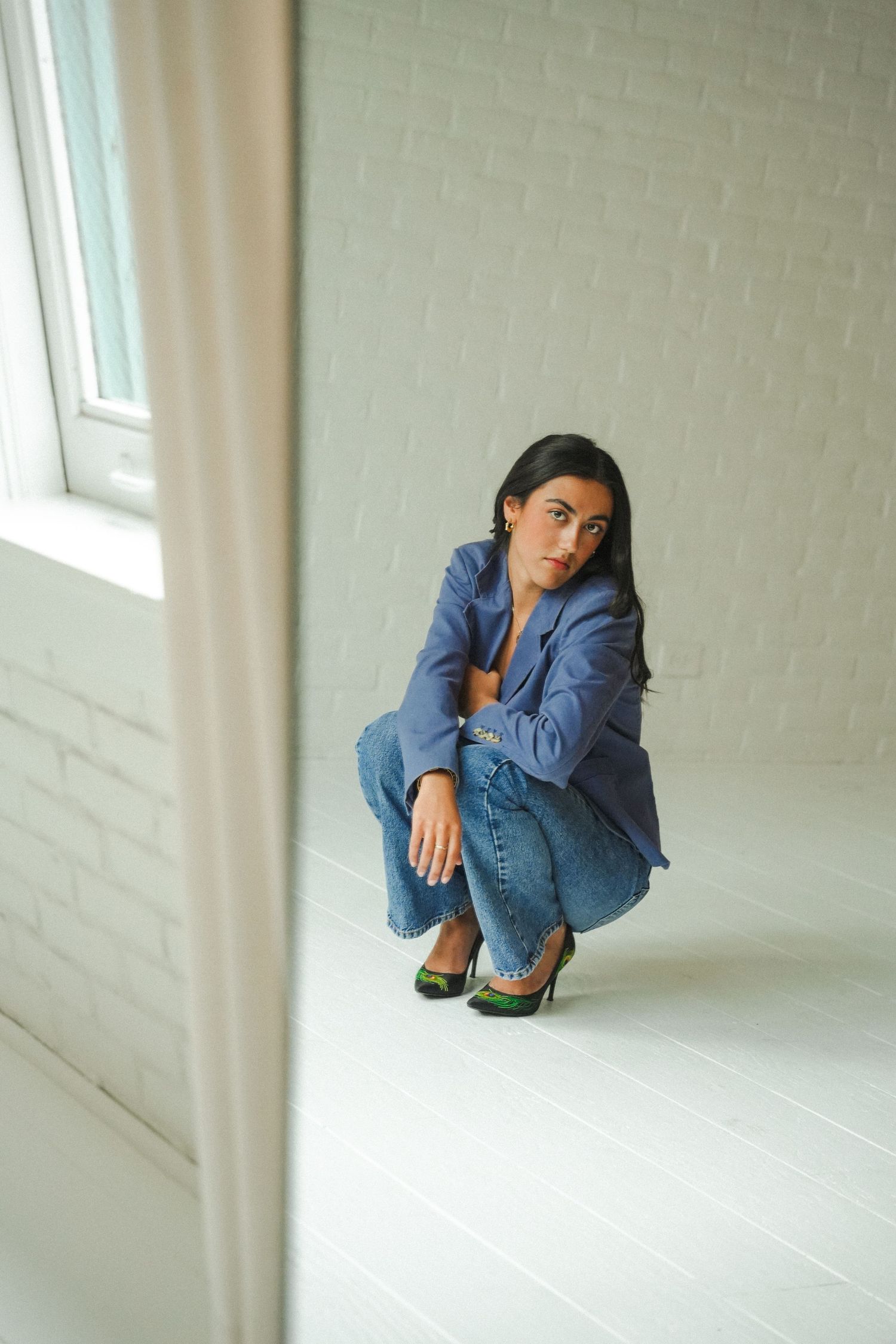Woman in blue blazer and jeans crouching against white brick wall near window in minimalist studio.