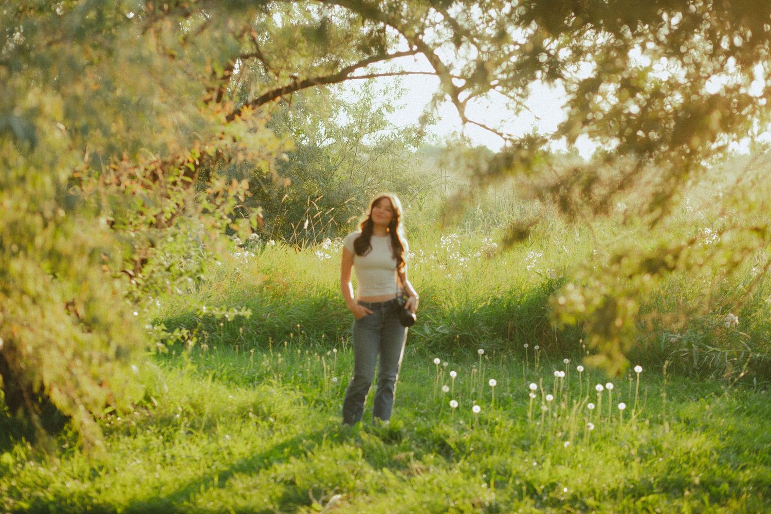 Young woman standing in a sunlit green meadow under tree branches, wearing jeans and white top.