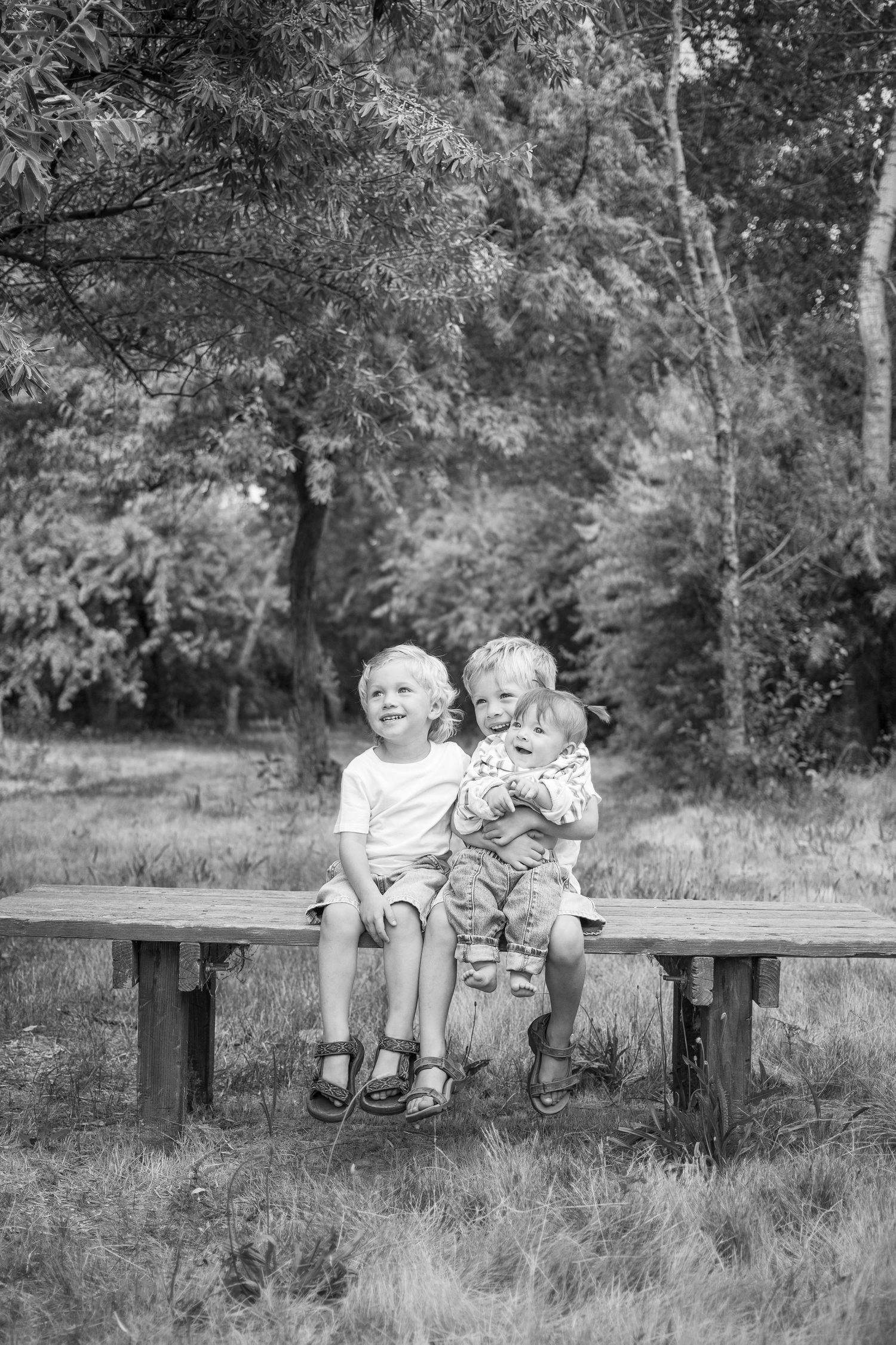 Two young children sit together on a wooden bench surrounded by lush trees in a black and white outdoor portrait.