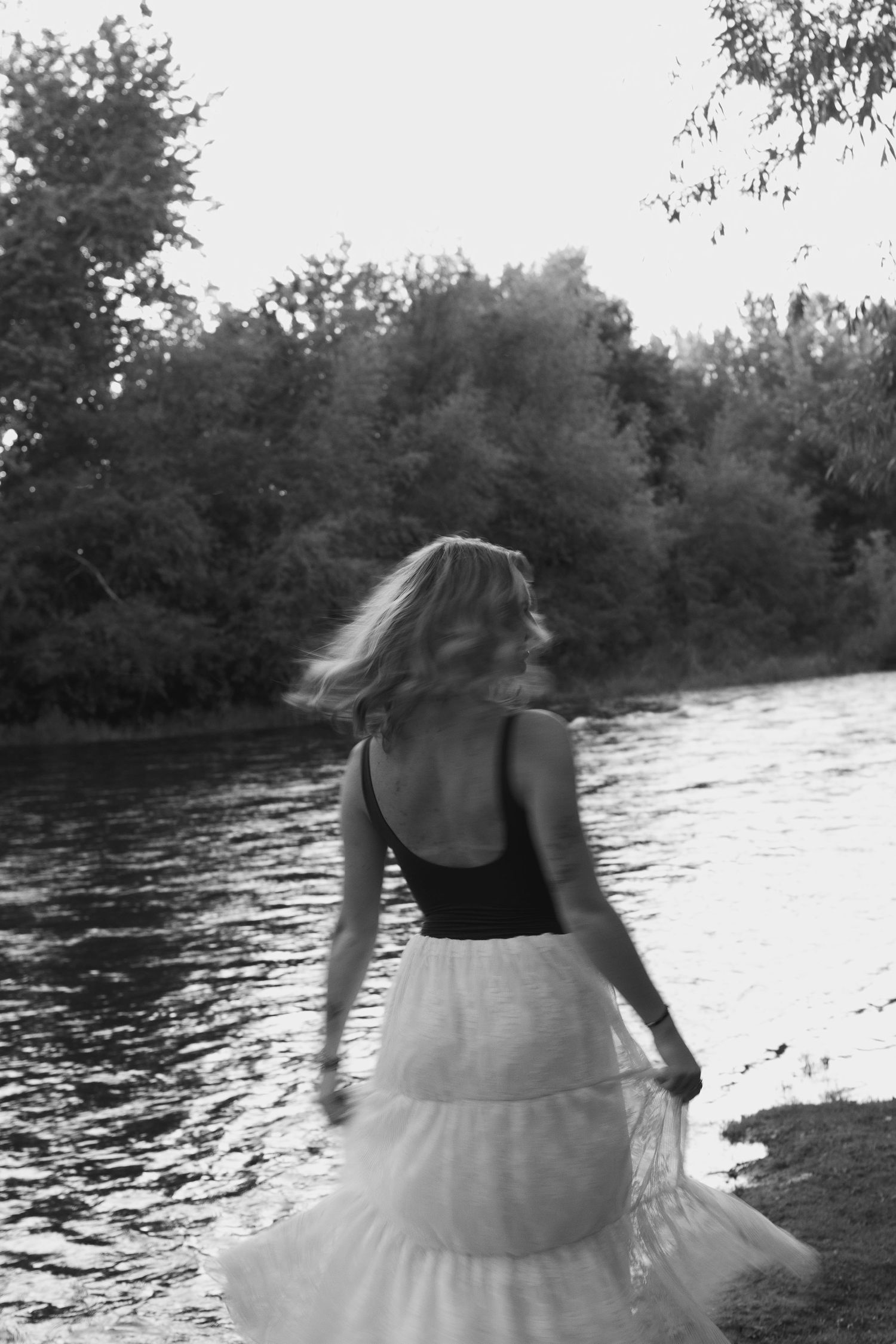 Black and white photo of a woman in a tank top and tulle skirt walking near a river with lush trees.