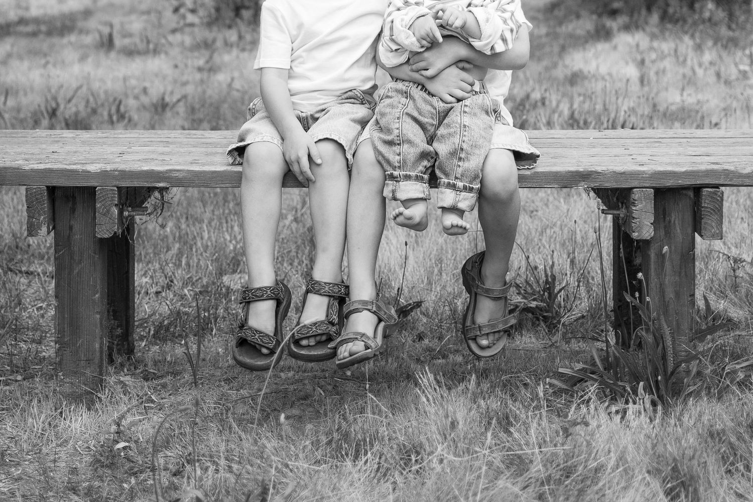 Black and white close-up of two children sitting on a wooden bench wearing sandals.
