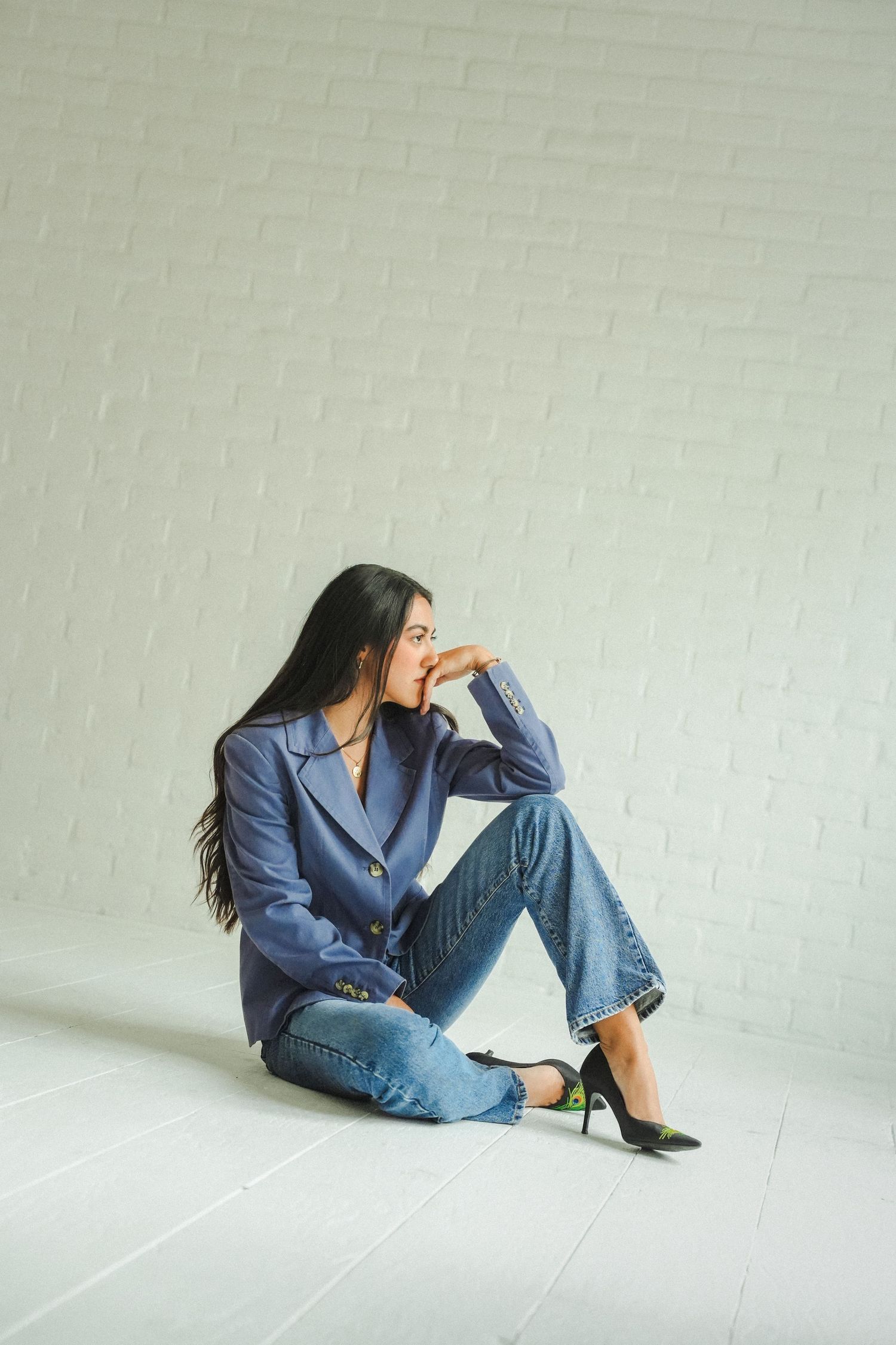 A stylish young woman in a blue blazer and jeans sits on a white floor against a brick wall, posing thoughtfully.