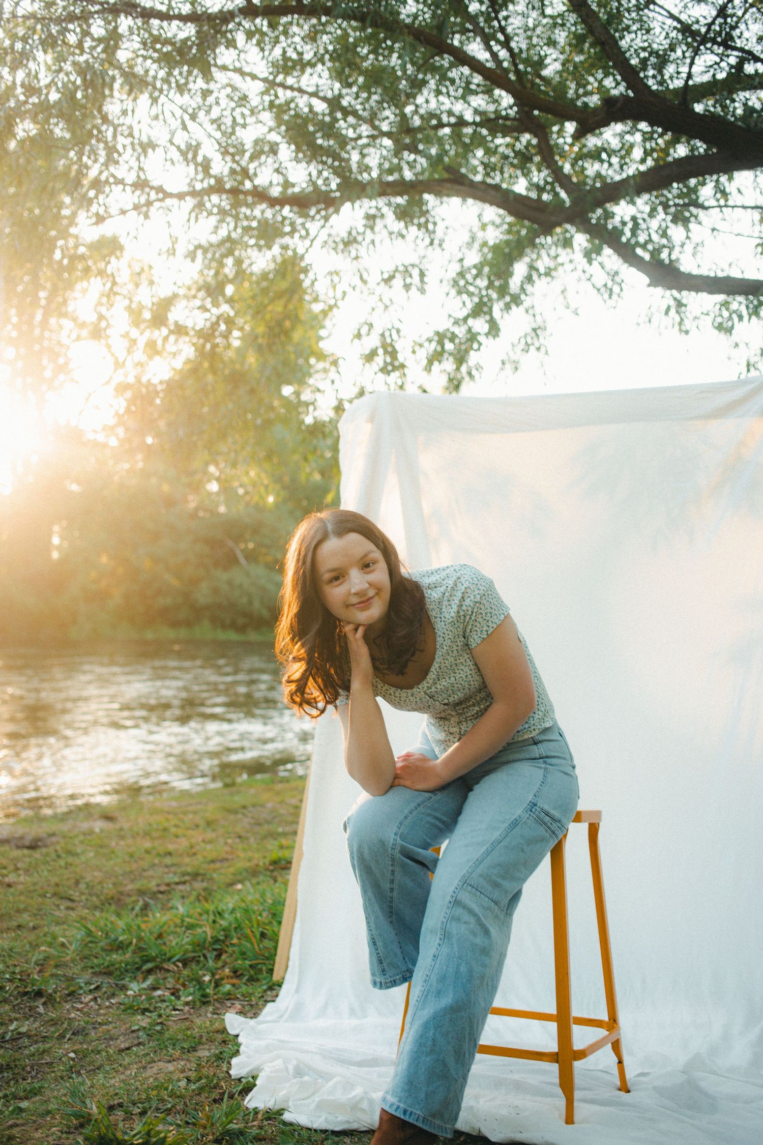 A smiling teenage girl sits on a yellow stool beside a white backdrop in golden sunset light outdoors.