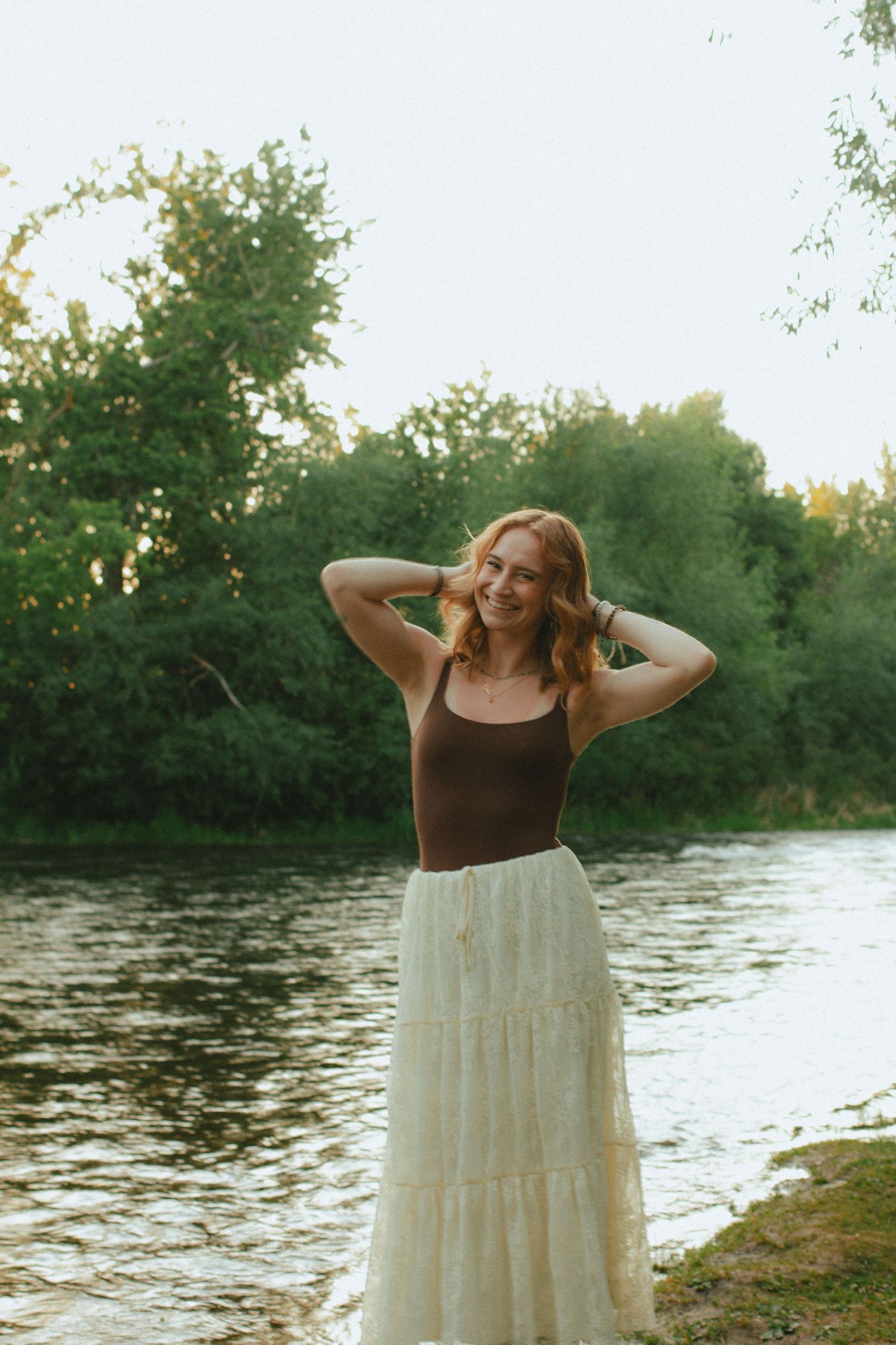 Smiling redhead woman in brown tank top and white lace maxi skirt posing by a river with green trees.