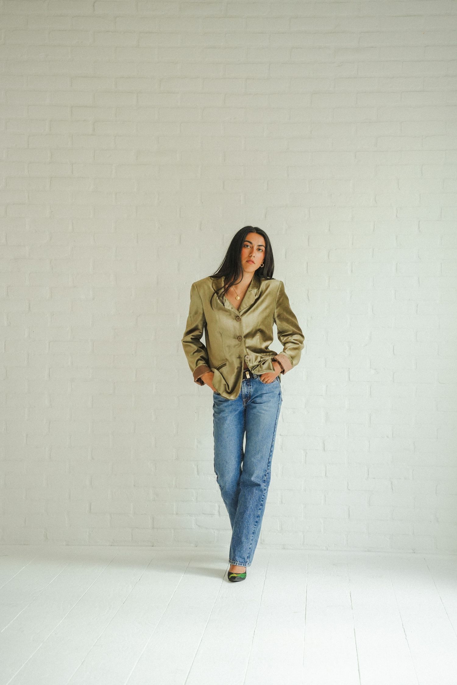 Woman in olive satin blazer and blue jeans posing confidently against a white brick wall.