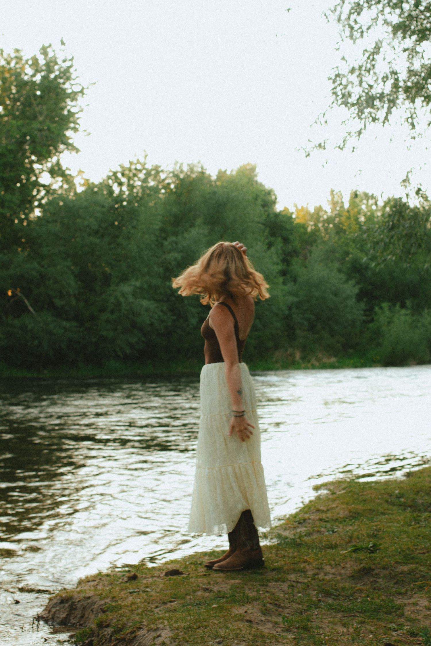 A blonde woman in a white maxi skirt and brown boots stands by a serene river at golden hour.