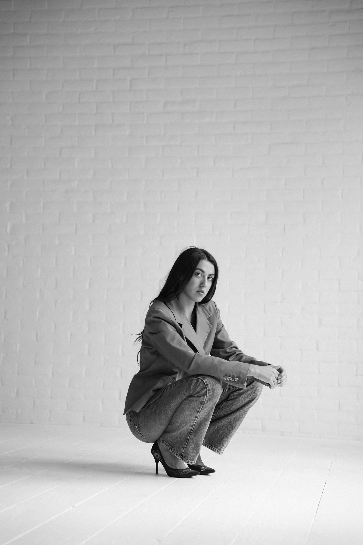 Black and white photo of a young woman crouching in jeans and heels against a white brick wall.