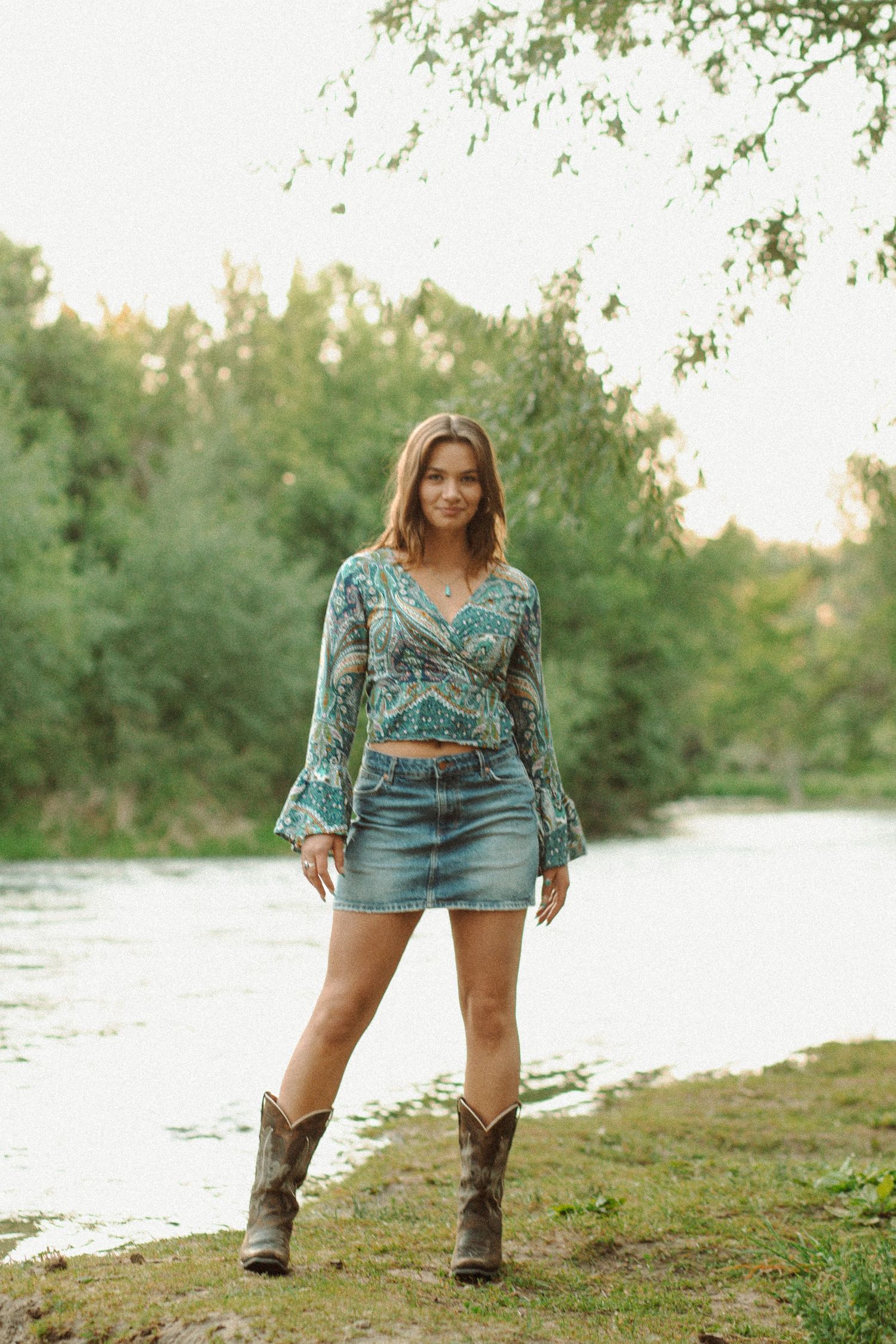 Woman in boho paisley top, denim mini skirt, and cowboy boots stands by a lush green riverbank.
