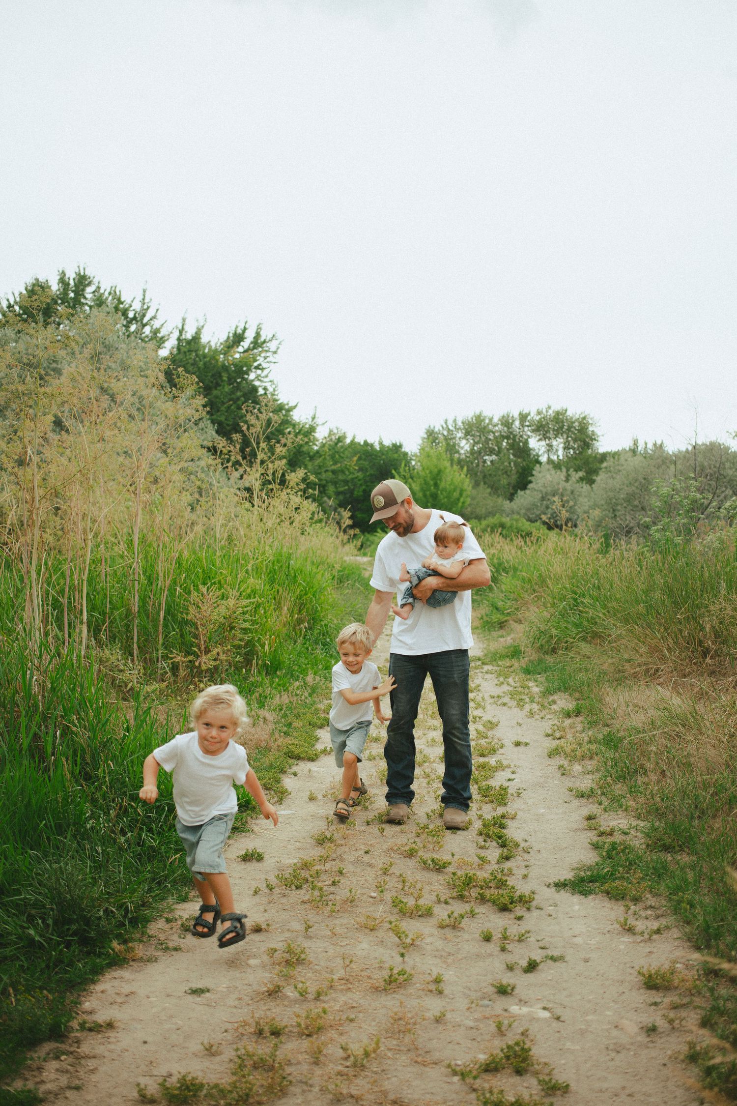 Father holds baby while two young boys run along a scenic nature trail surrounded by tall grass.