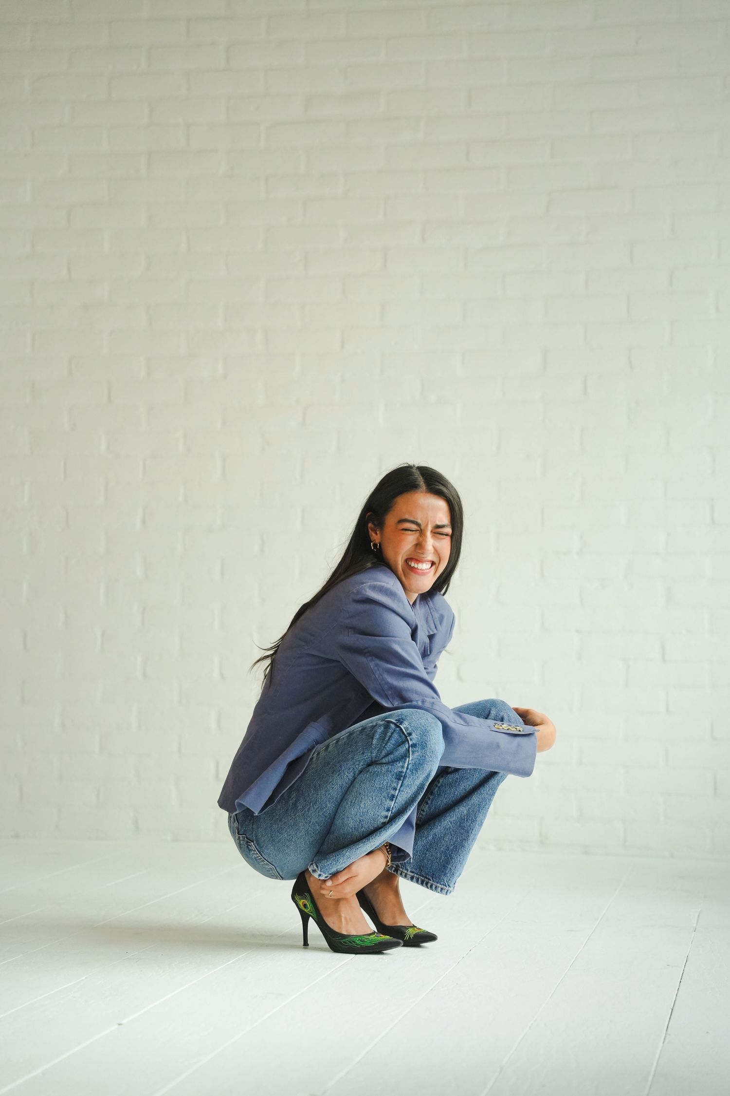 A smiling woman in a blue blazer and jeans crouches playfully in front of a white brick wall.