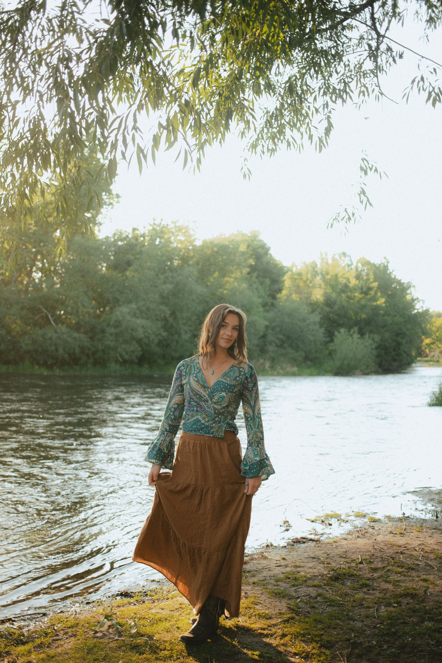 Woman in paisley boho top and brown maxi skirt stands gracefully by a sunlit green riverbank.