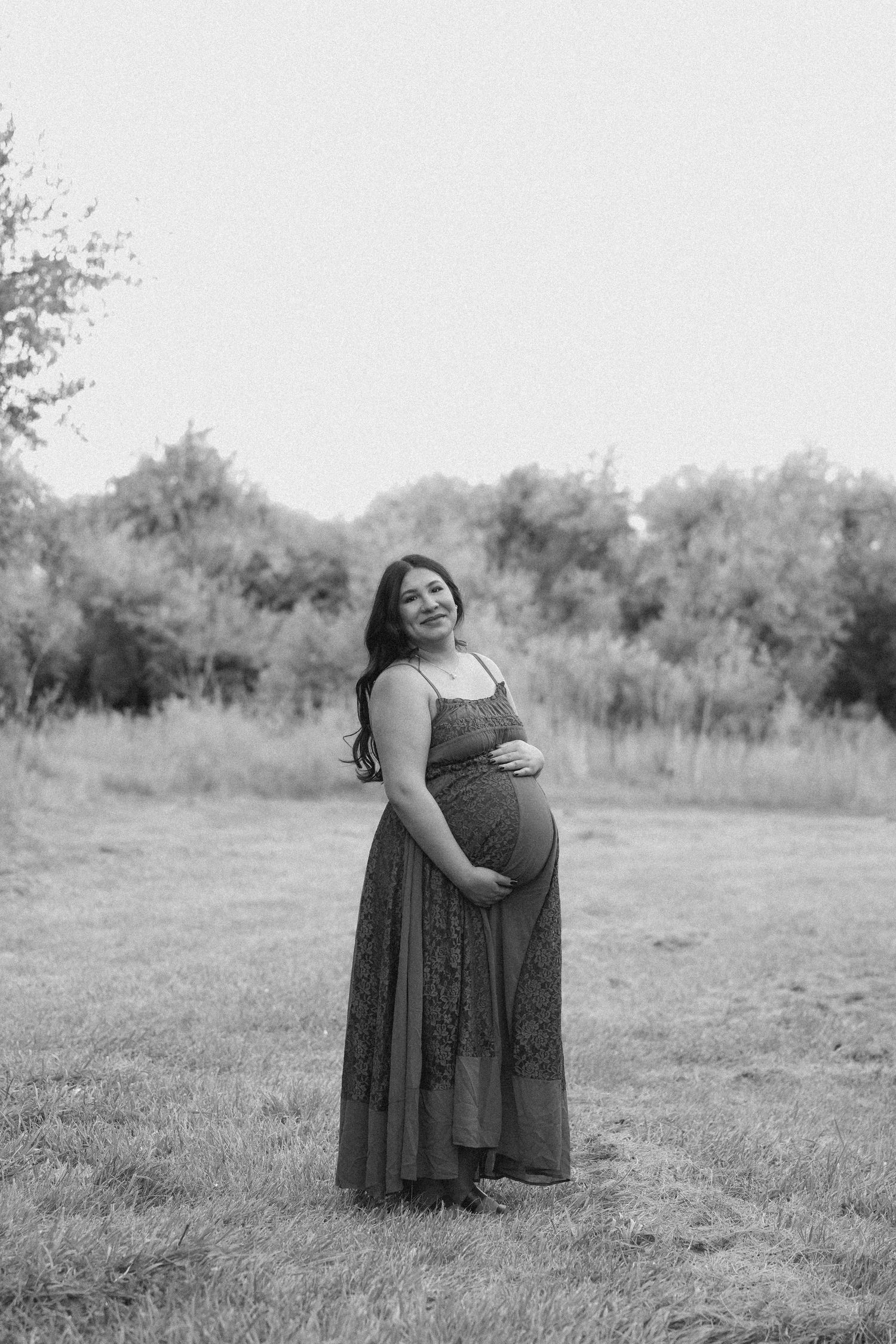 Black and white maternity photo of a smiling pregnant woman in a maxi dress holding her belly in a grassy outdoor field.
