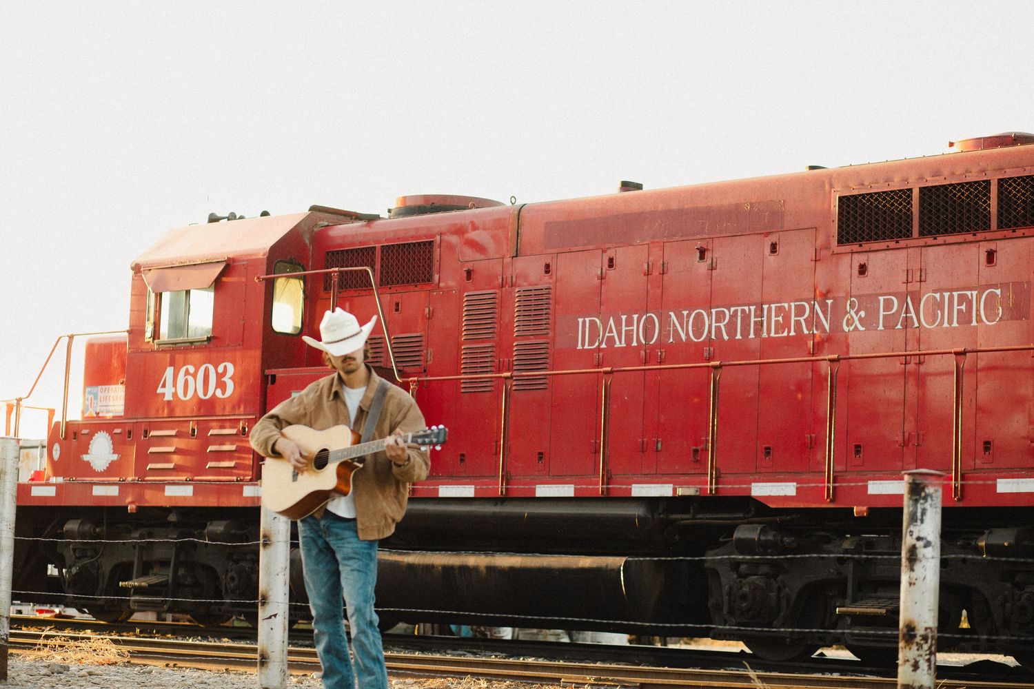 Cowboy musician playing acoustic guitar in front of red Idaho Northern & Pacific locomotive 4603 at railroad yard.