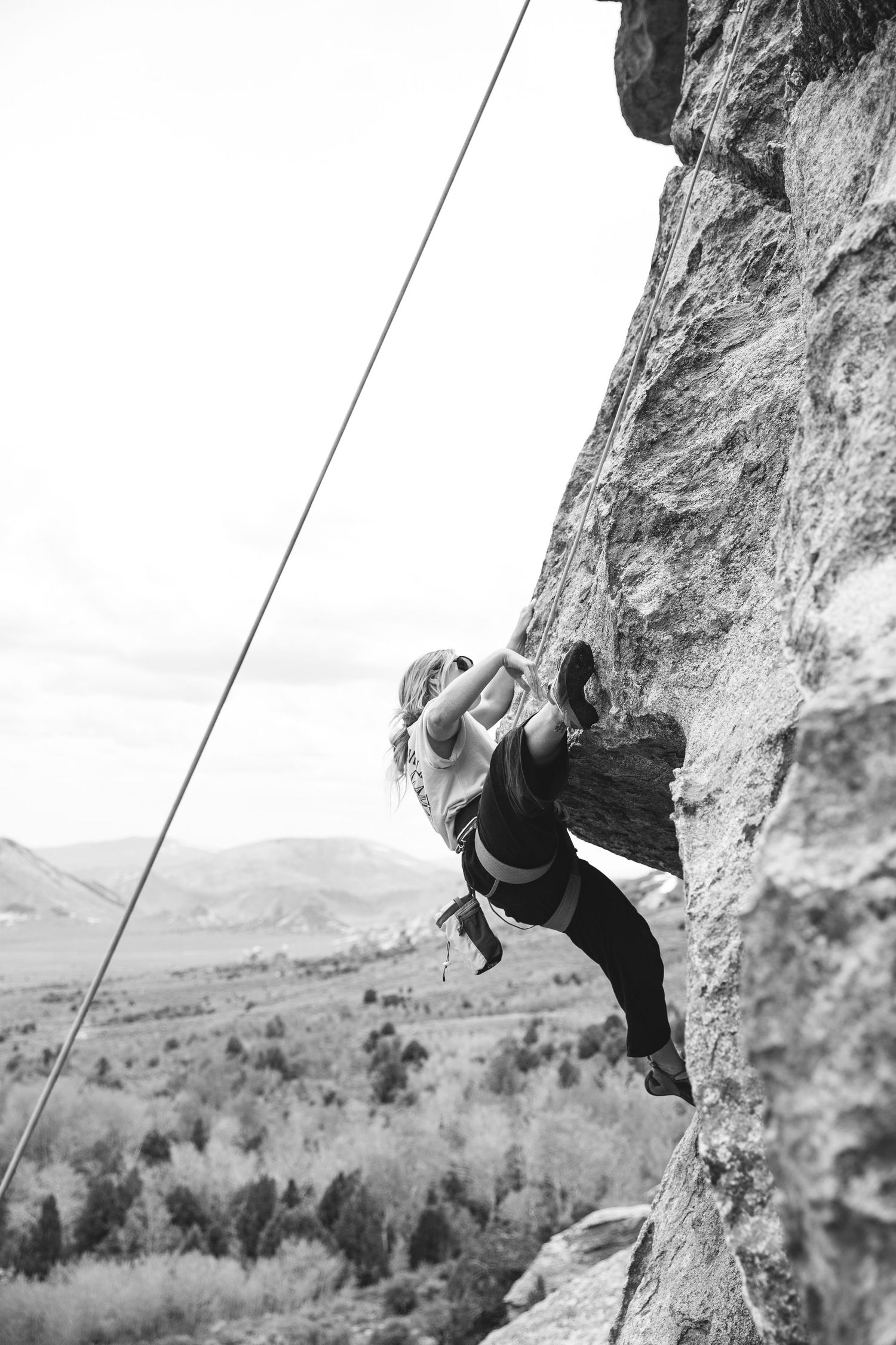 A rock climber scales a steep cliff face while secured by ropes, with a scenic mountain valley landscape in the background.