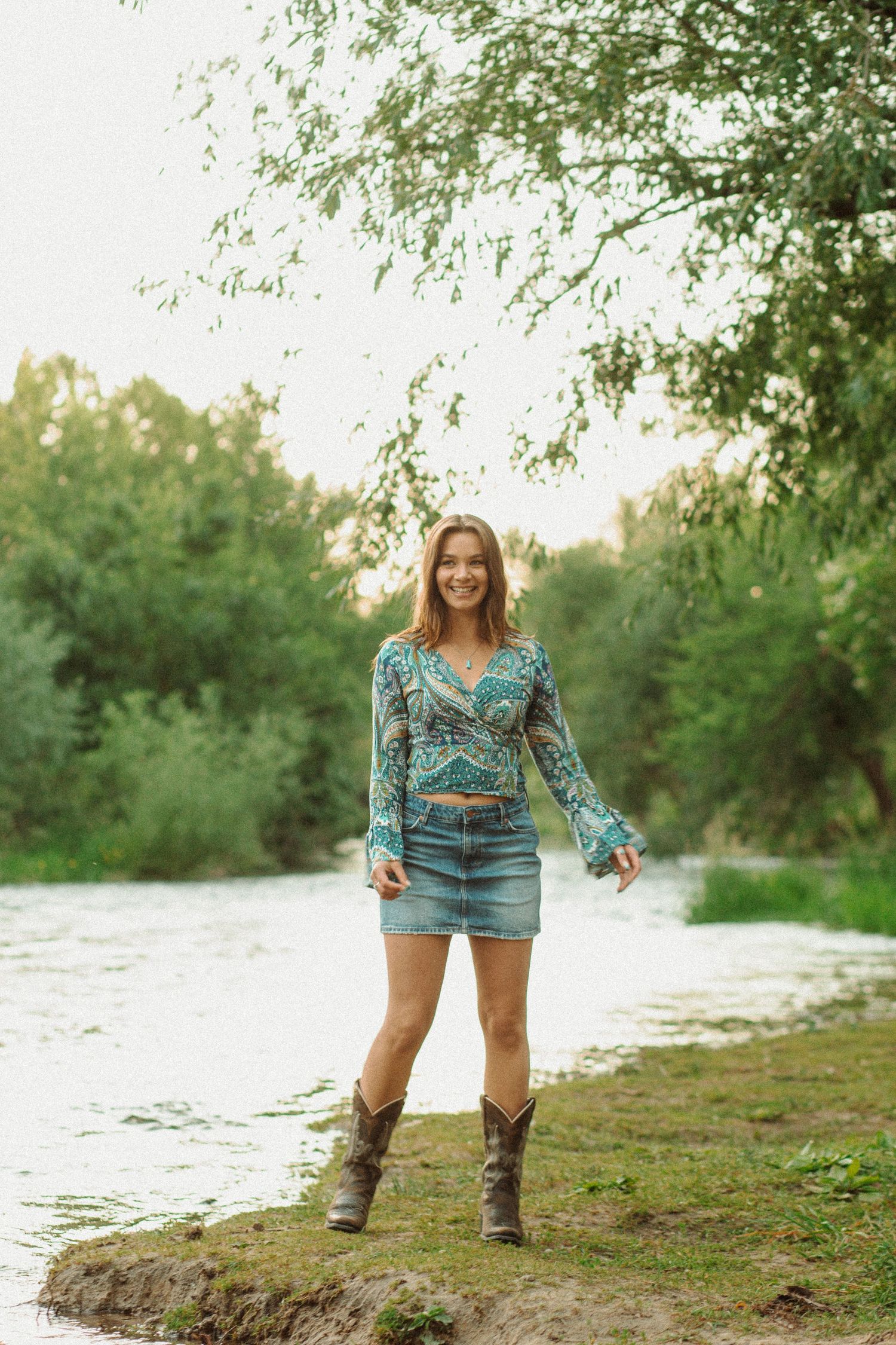Smiling woman in teal paisley top, denim mini skirt, and cowboy boots standing by a riverside.