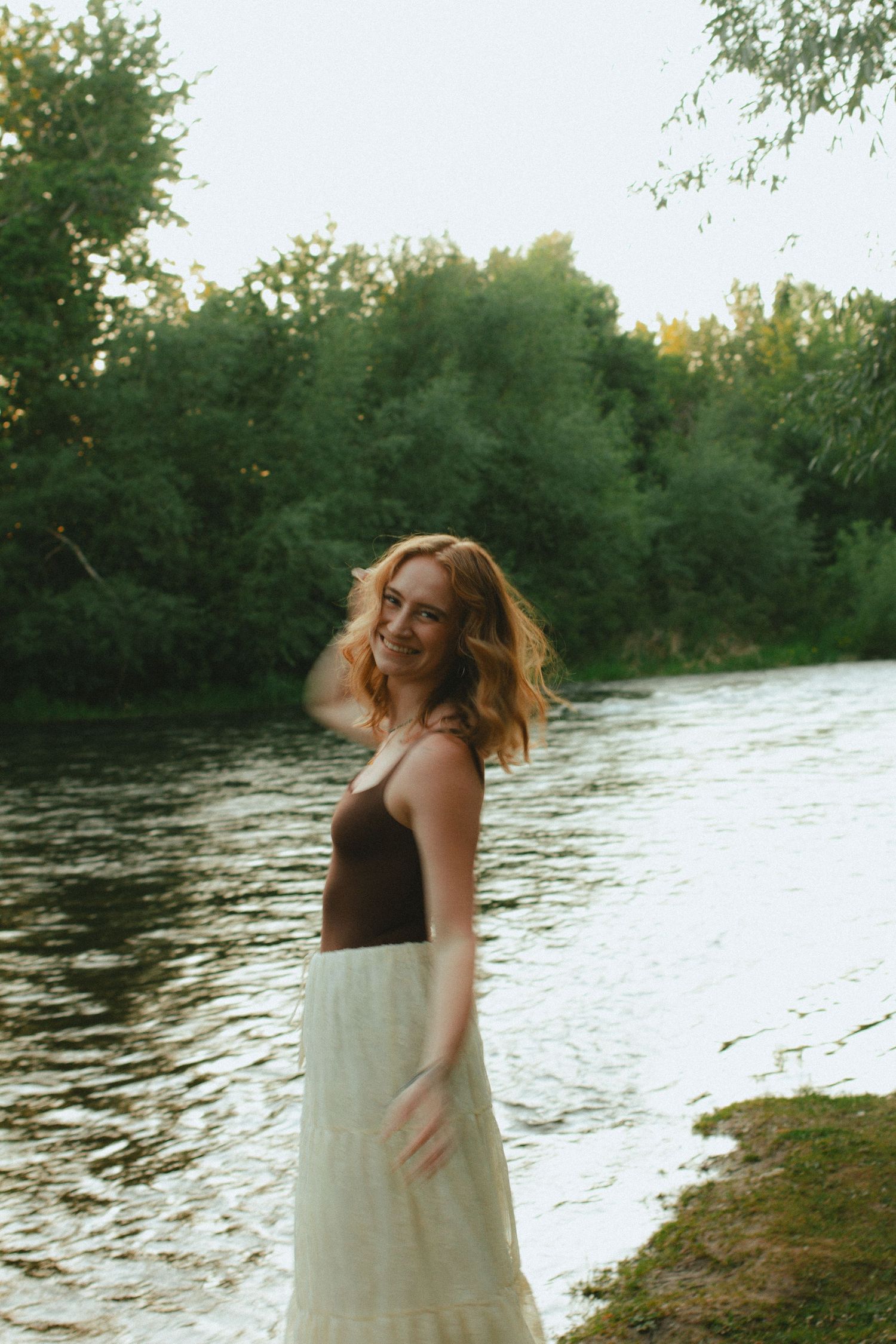 A smiling young woman in a white skirt stands by a peaceful river surrounded by lush green trees at dusk.