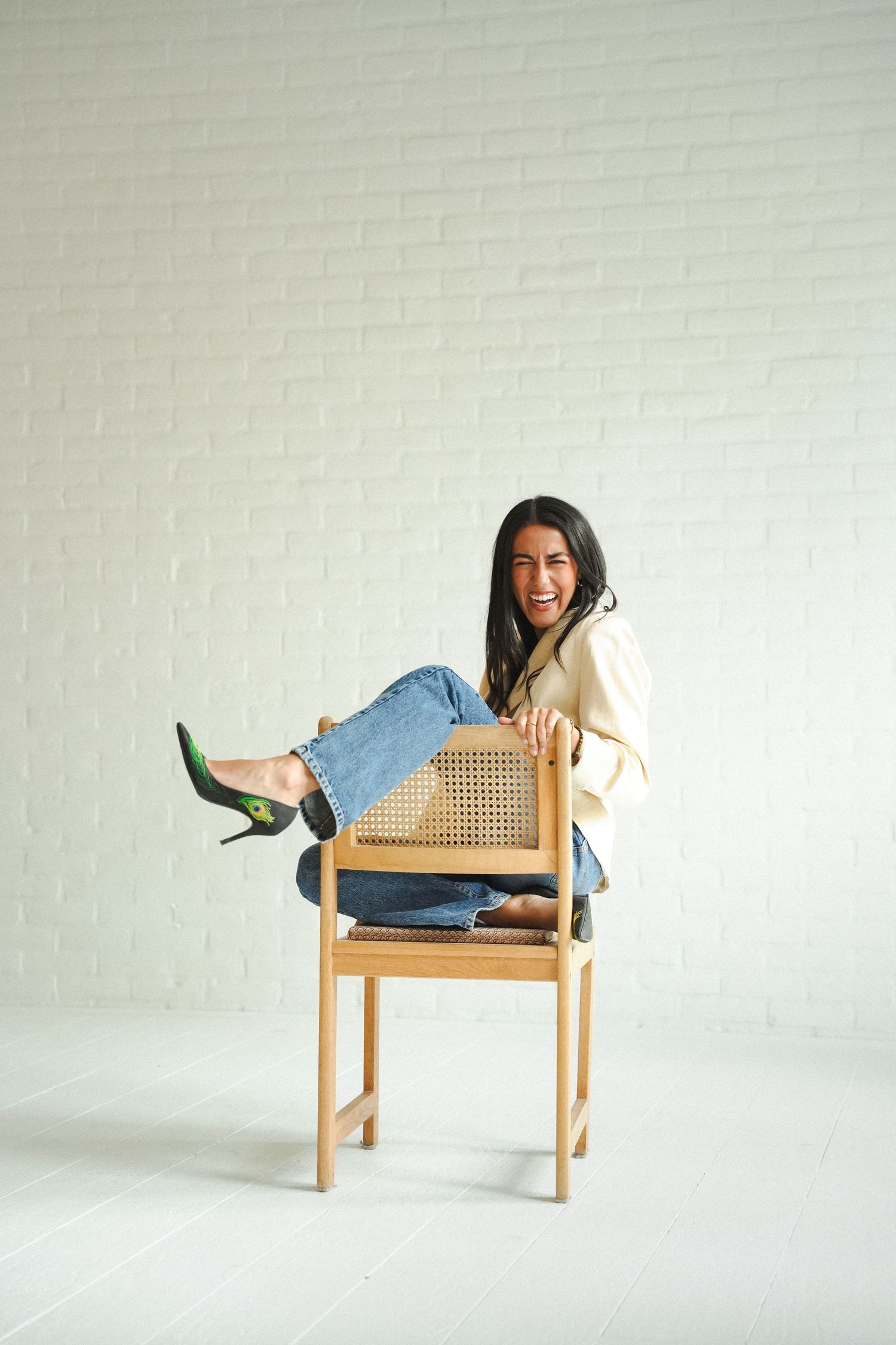 Woman in jeans and blazer sitting playfully on wooden rattan chair against white brick wall, laughing.