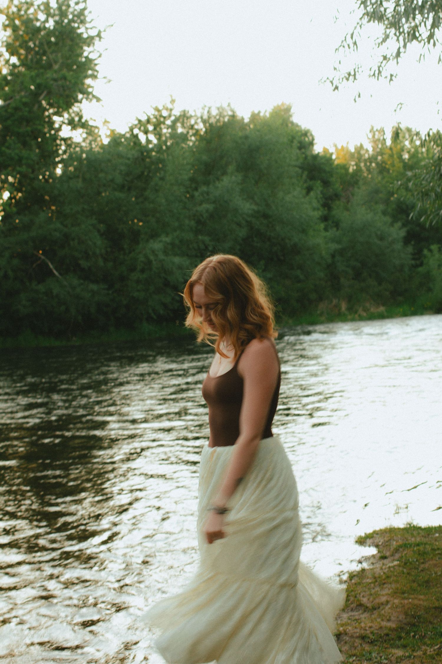 Young woman in brown tank top and white tulle skirt walking near a calm river surrounded by green trees.