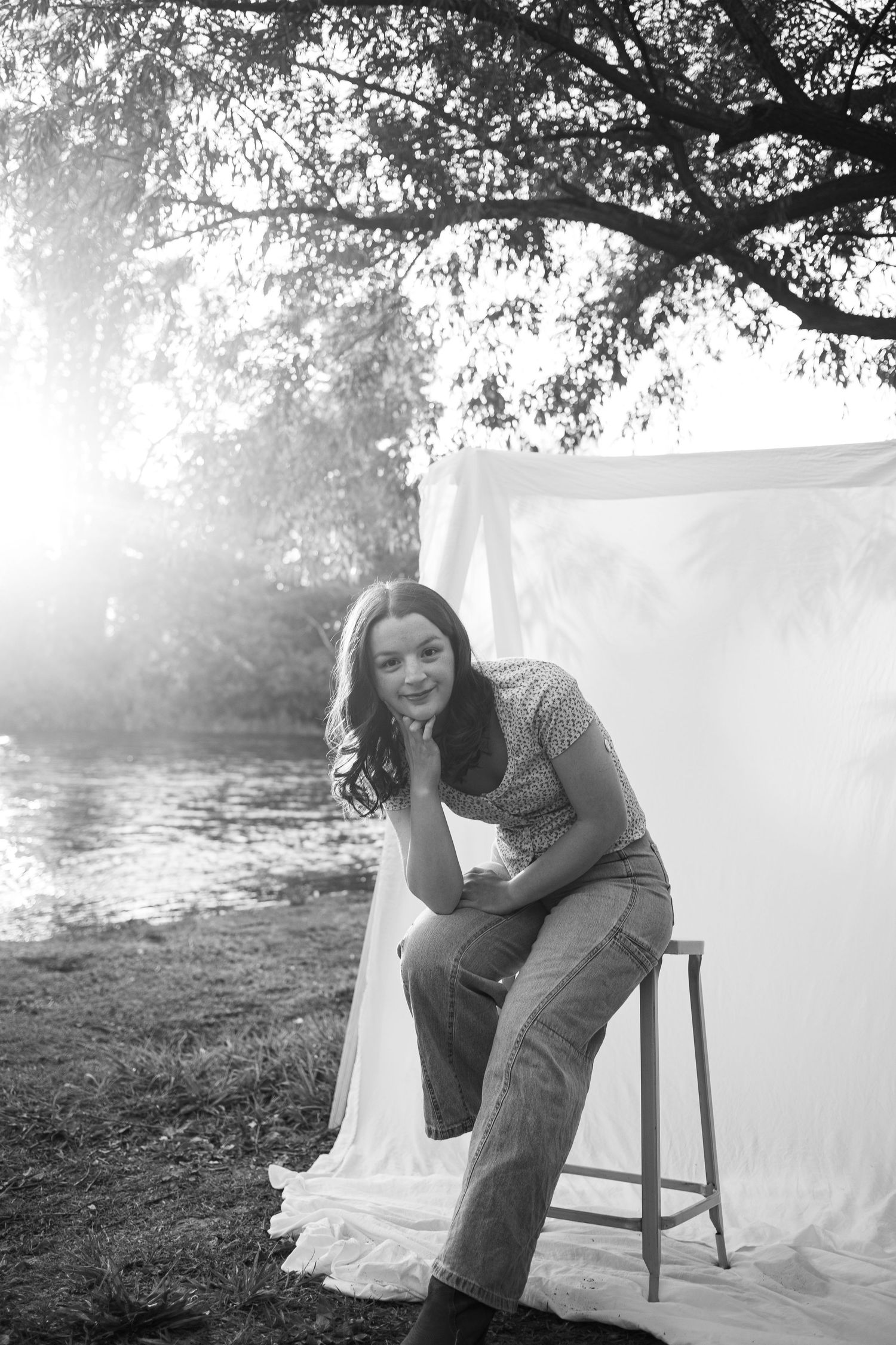 A young woman sits on a stool beside a white backdrop near a sunlit river in black and white.