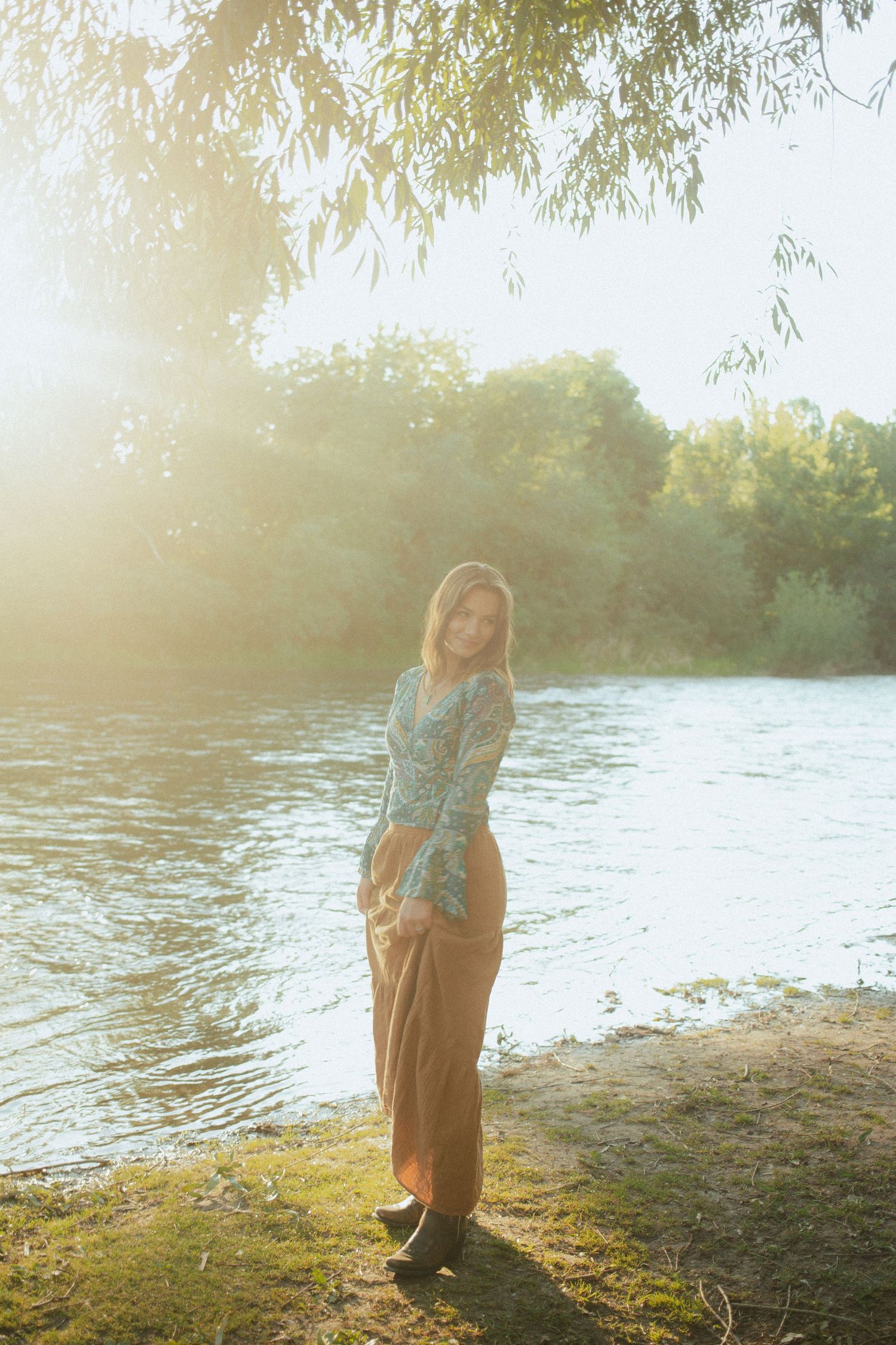Young woman in floral blue top and brown wide-leg pants standing by a sunlit river surrounded by trees.