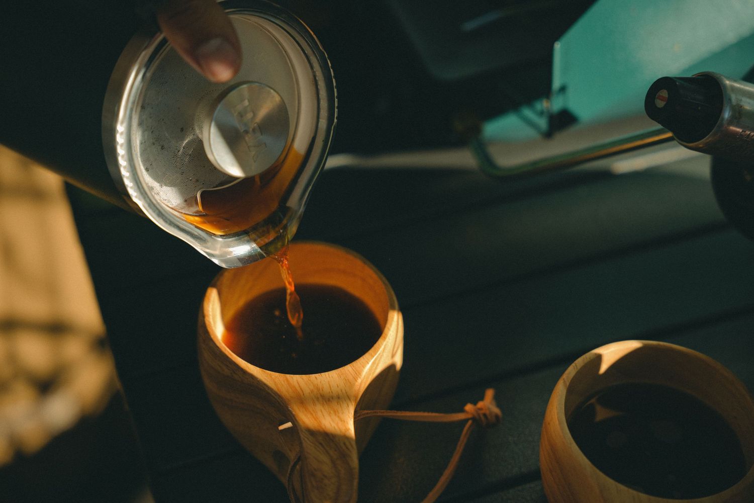 Person pouring hot coffee from a glass kettle into a wooden cup in a dimly lit car interior.