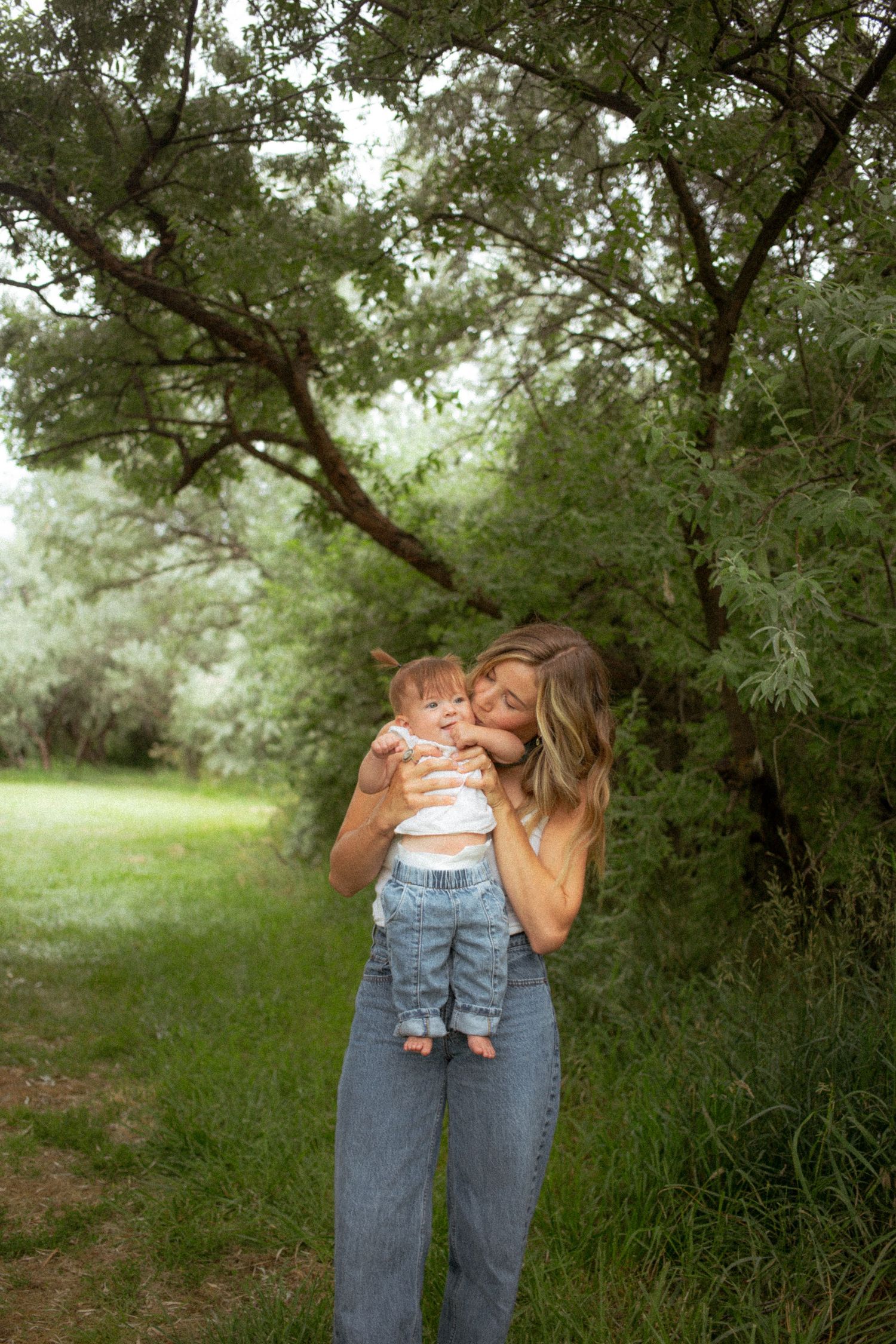 Mother tenderly kissing her baby while standing in a green wooded area.
