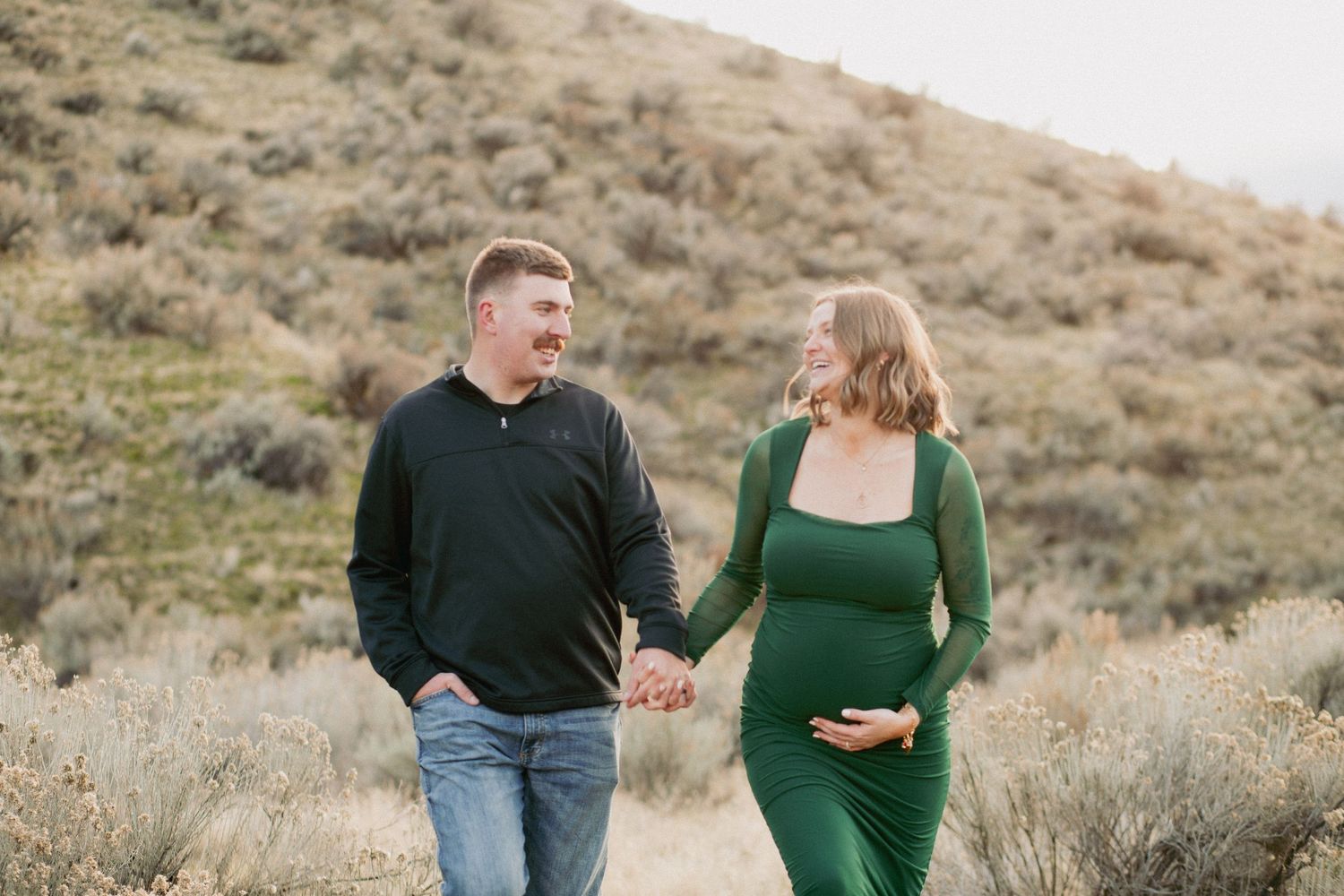 Pregnant couple holding hands and smiling outdoors in a field with green hills, woman in green dress cradling baby bump.