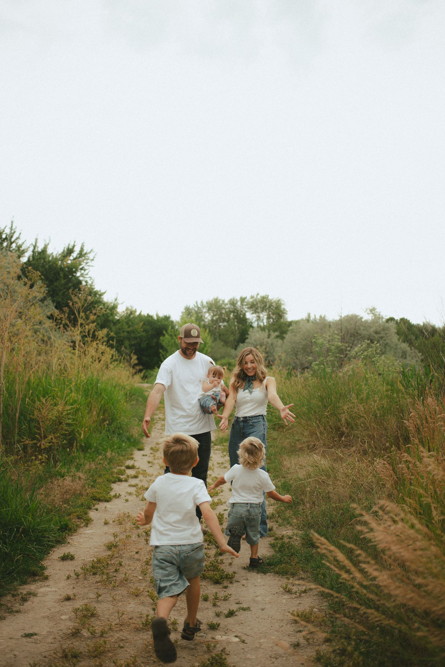 A young family walks along a scenic dirt path through tall grasses in a warm outdoor nature setting.