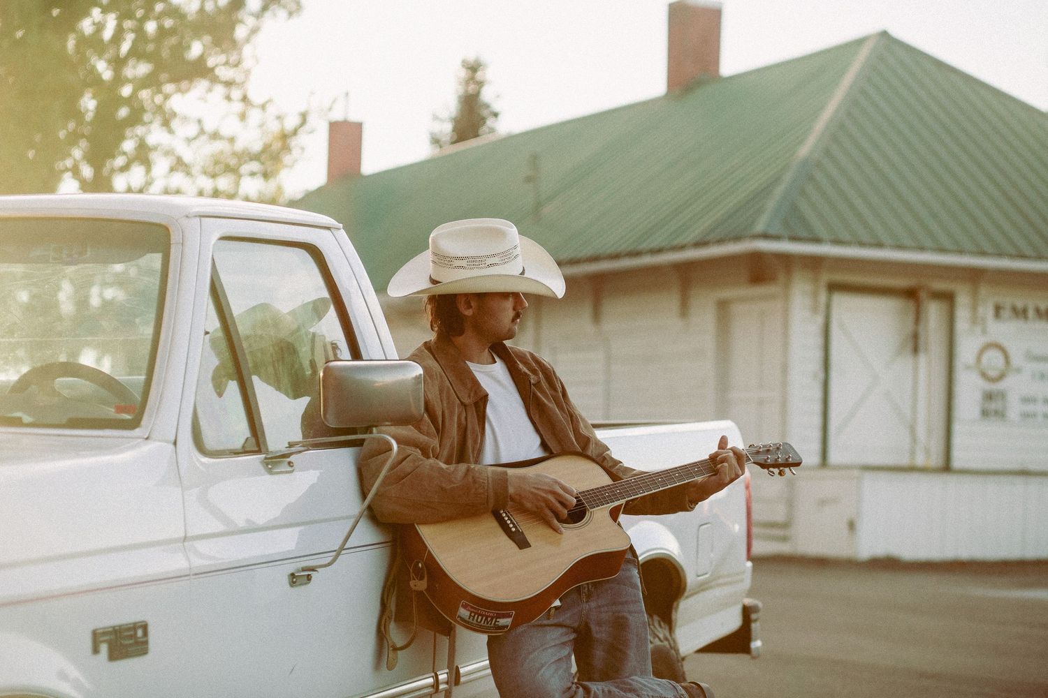 A cowboy musician plays acoustic guitar leaning against a white truck near a rustic building in warm golden sunlight.