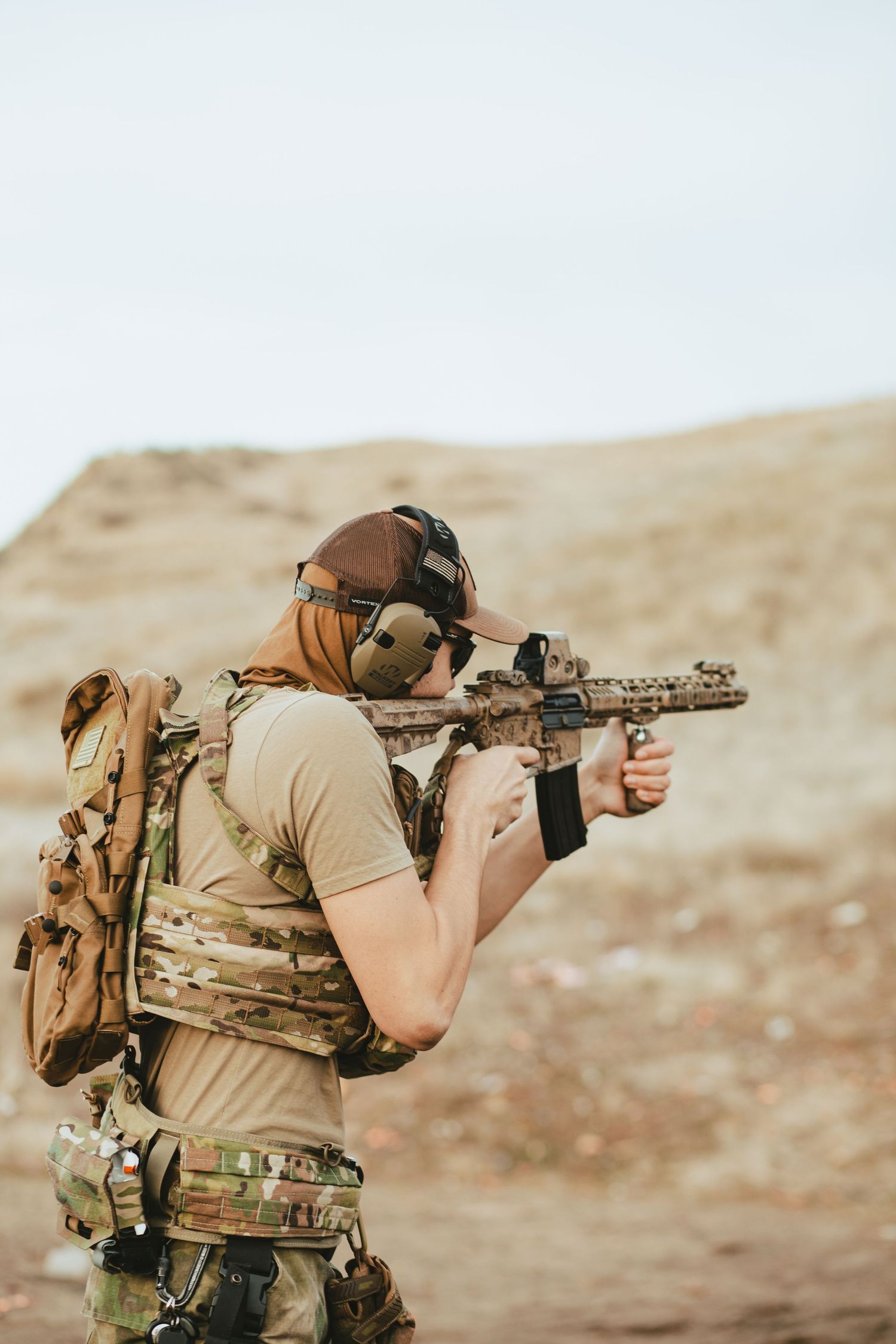 Tactical shooter in desert camouflage gear aims rifle while wearing backpack in arid environment.