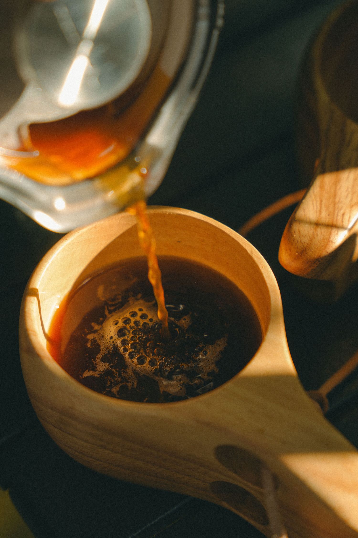 Hot coffee being poured from a glass kettle into a wooden cup, illuminated by warm sunlight.